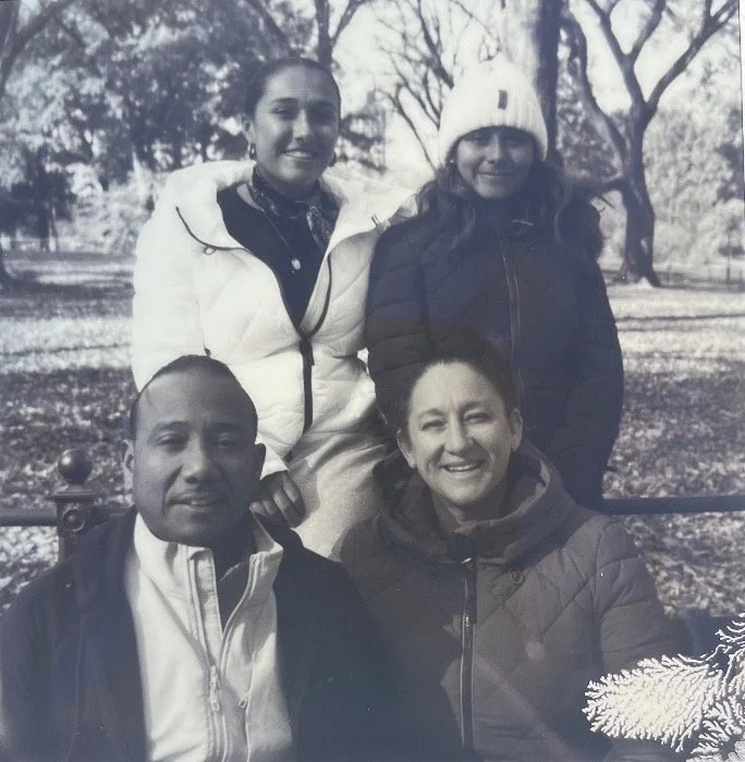 A black and white photo of two adults and two children outdoors in a park with trees. The adults are seated, and the children are standing behind them. The woman is smiling, the man has a neutral expression, and the children are smiling.