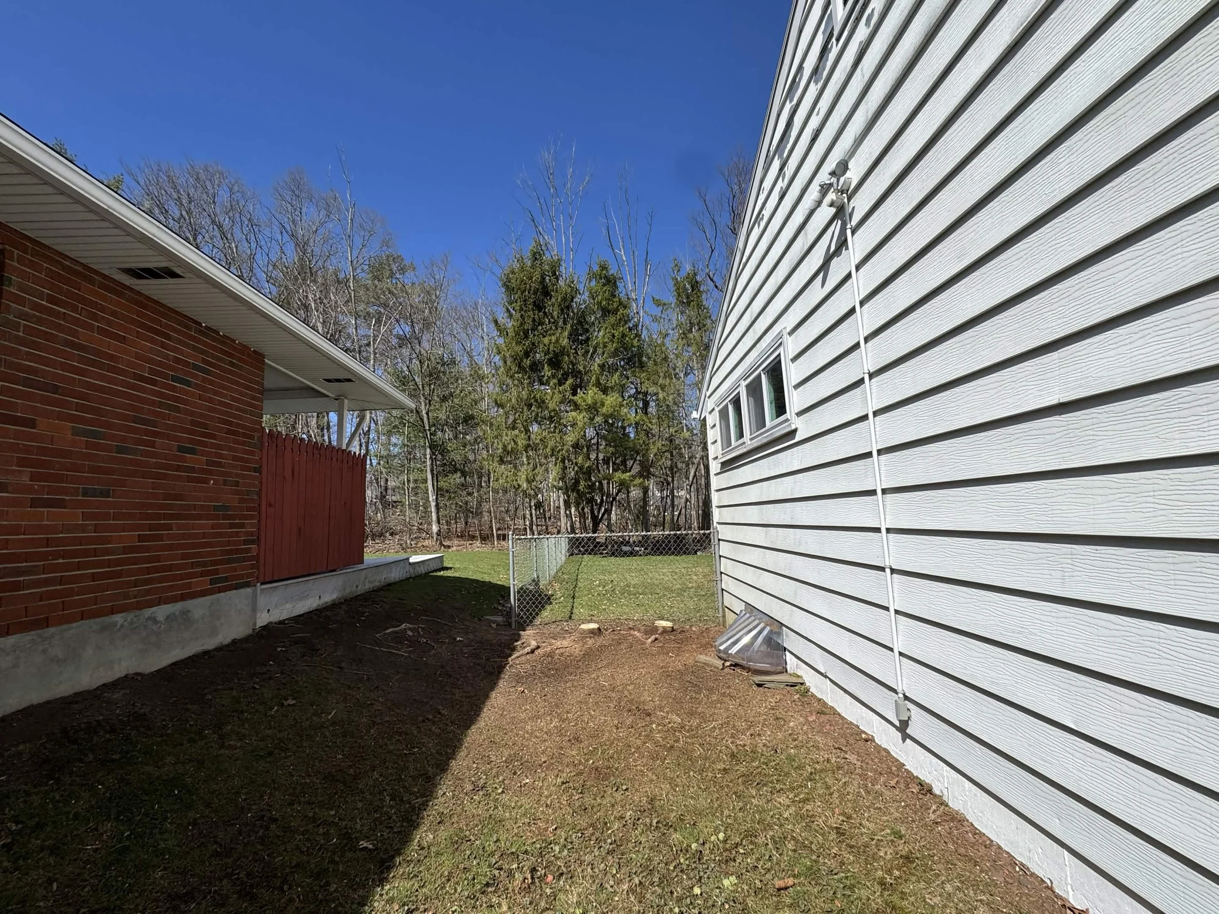 Exterior view of two houses with a dirt backyard, a chain-link fence, and a row of trees in the background. One house has red brick walls; the other has white horizontal siding and a small window.