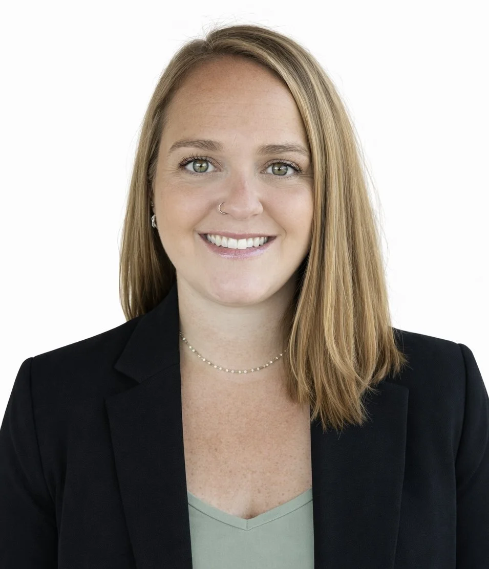 Portrait of a young woman with shoulder-length red hair, wearing a black blazer and a light green top, smiling at the camera against a white background.