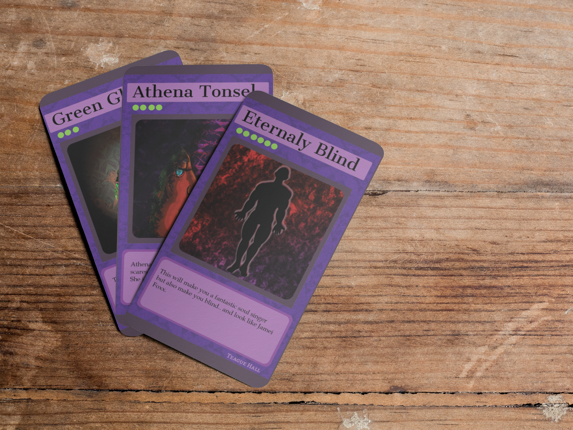 Three purple cards with fantasy-themed artwork and text, spread out on a wooden surface. The cards feature titles: 'Green G', 'Athena Tonse!', and 'Eternally Blind'.