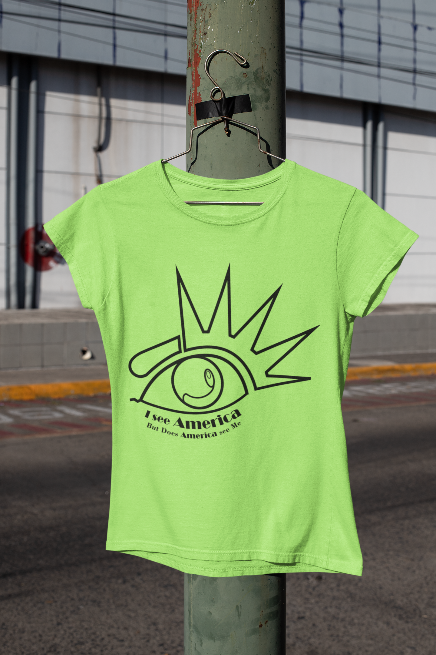 A bright green T-shirt hanging on a wire hanger tied to a green metal pole with red paint marks. The T-shirt has a graphic of a stylized eye with radiating lines and text that reads "I see America But Does America see Me". In the background, there is an urban setting with a building, power lines, and a sidewalk.