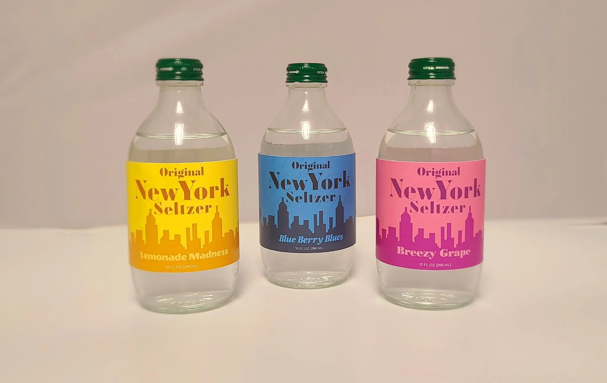 Three small bottles of flavored seltzer water on a white surface against a white background. The bottles are labeled 'New York Seltzer' with different flavor names: Lemonade Madness (yellow label), Blue Berry Blues (blue label), and Breezy Grape (pin
