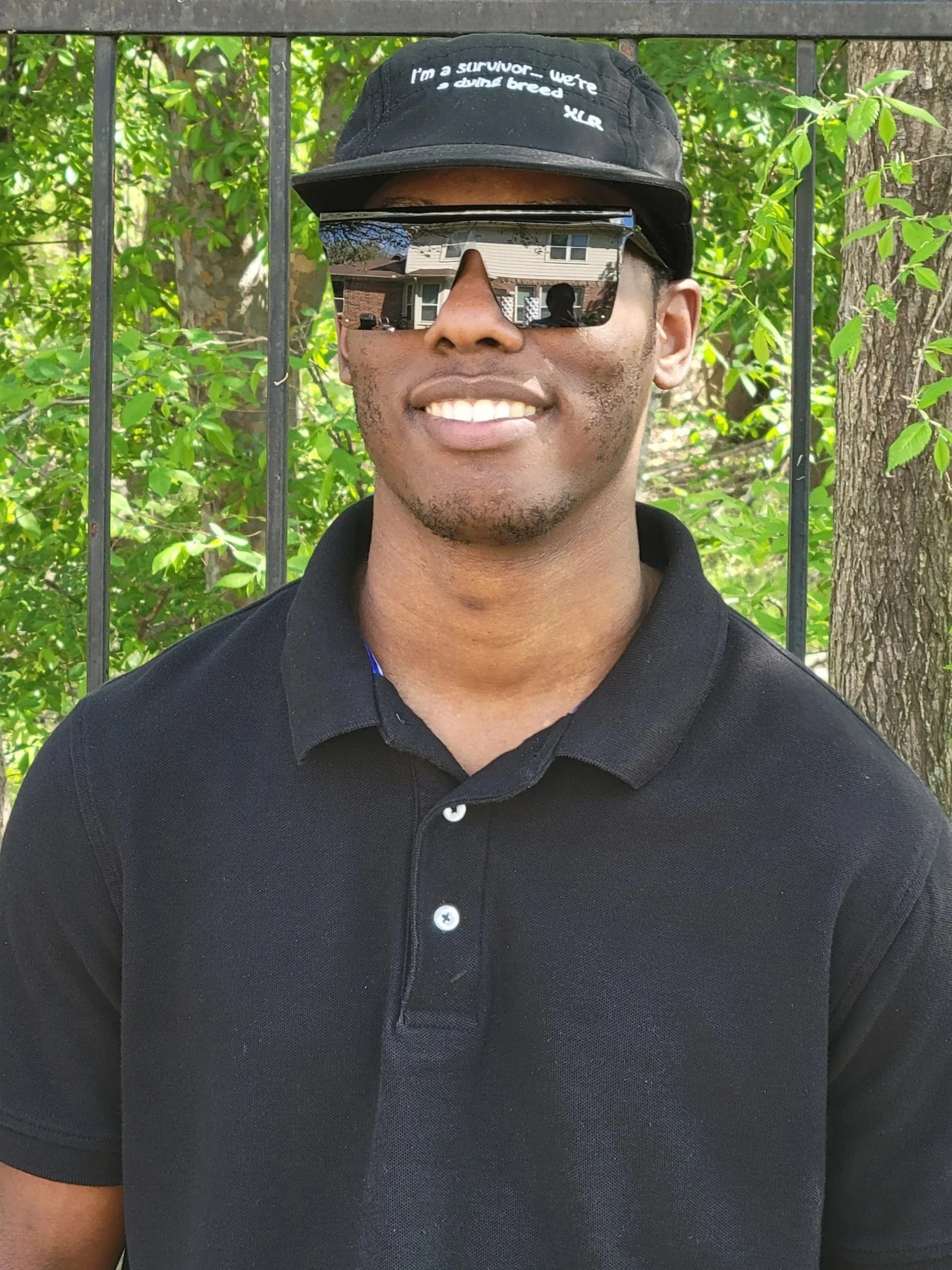 Close-up of a young man with a short beard, wearing a black baseball cap with a caption, standing outdoors behind a metal fence with tree leaves in the background. He is also wearing reflective sunglasses and a black polo shirt.