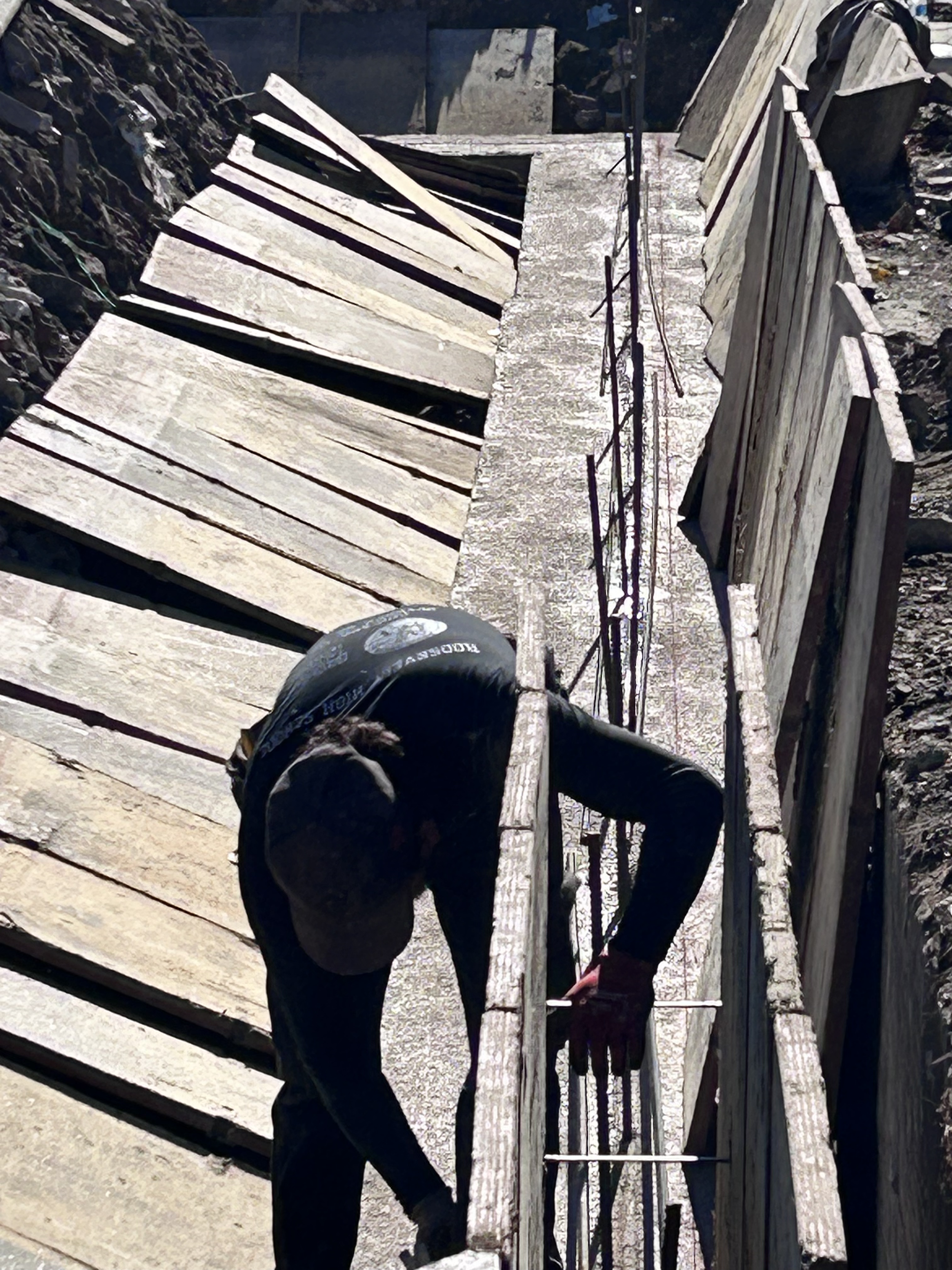 A construction worker wearing a black hoodie and baseball cap working on a concrete form for a sidewalk or pathway at a construction site.