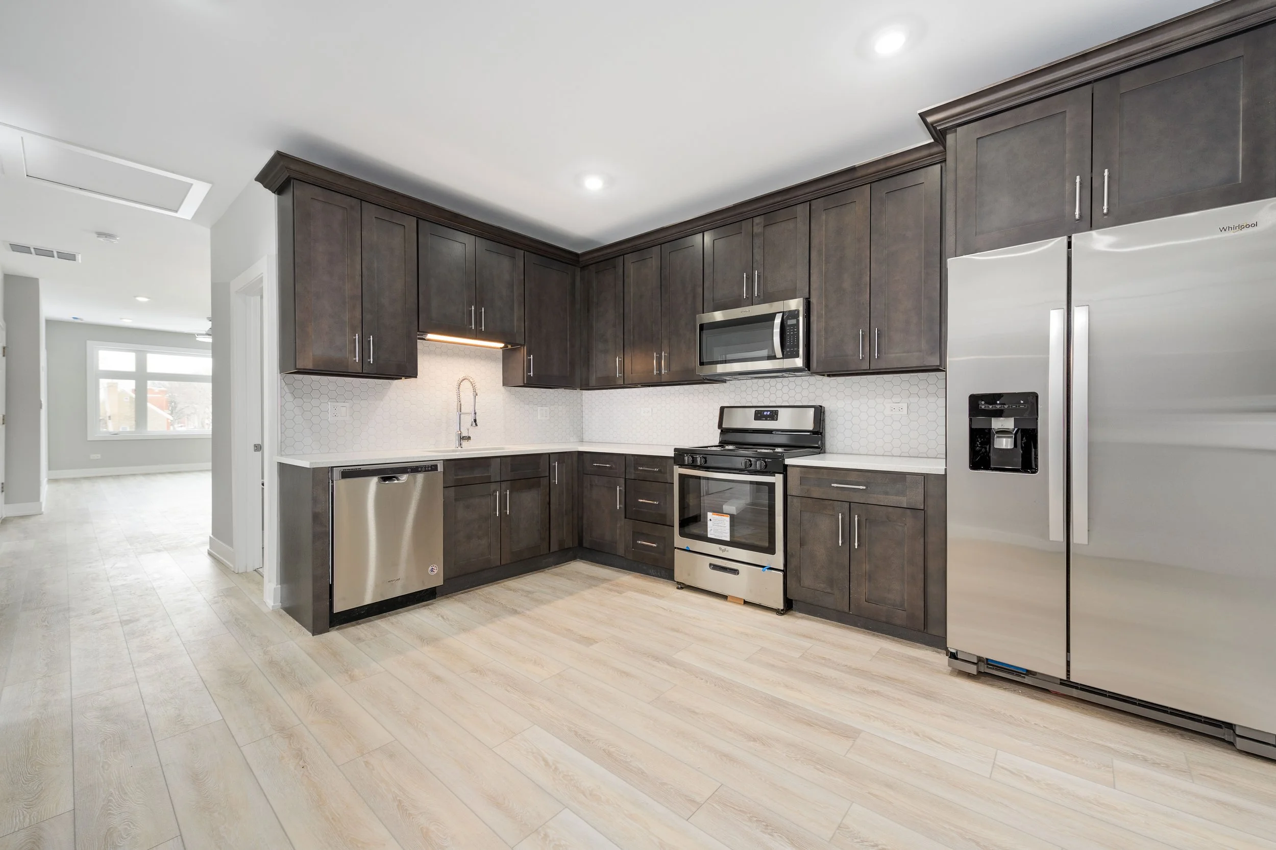 Modern kitchen with dark wood cabinets, stainless steel appliances, light wood floors, and a white hexagonal tile backsplash.