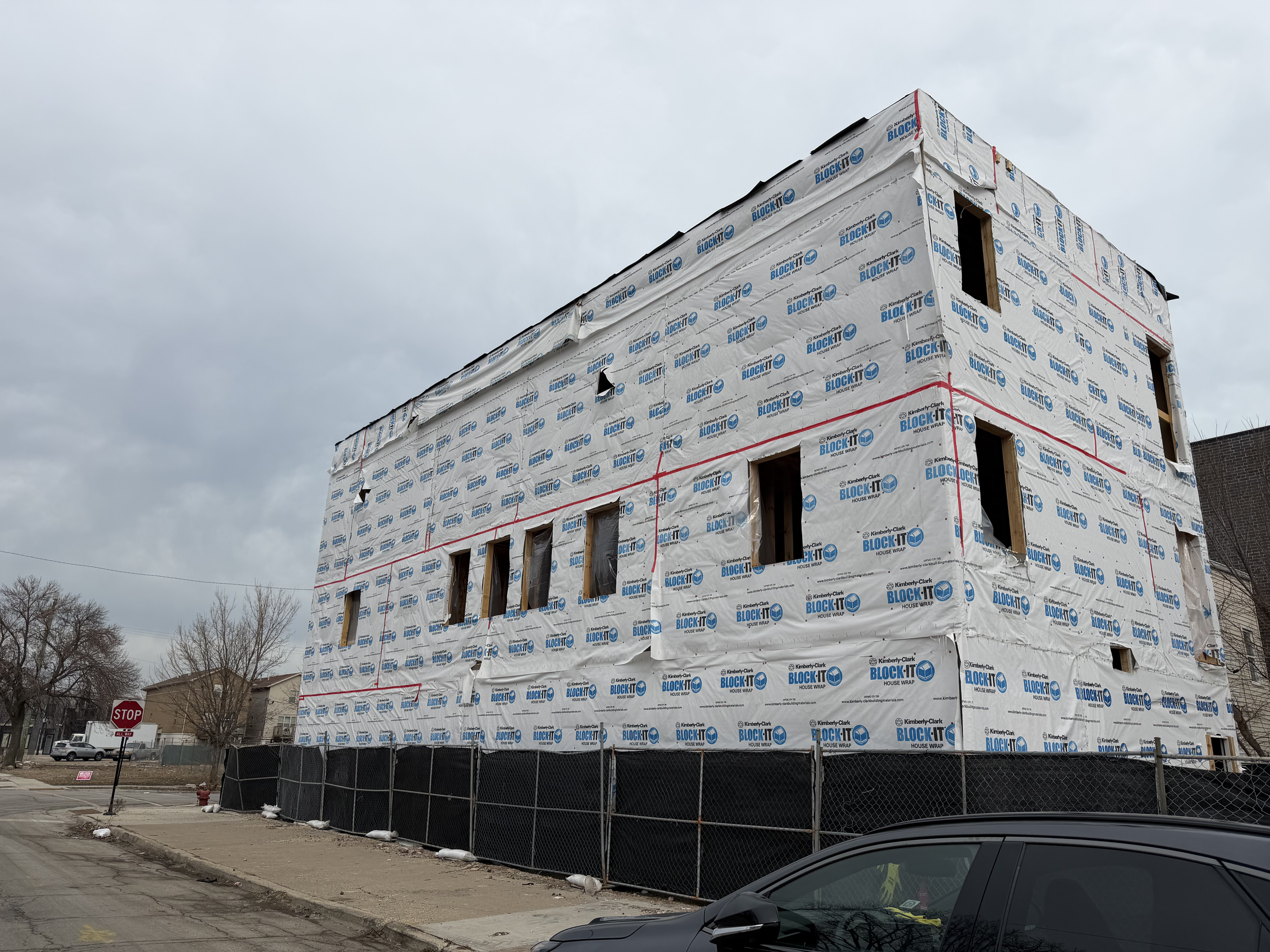 A three-story building under construction wrapped in white building paper with blue lettering, surrounded by a black construction fence, with a cloudy sky overhead.