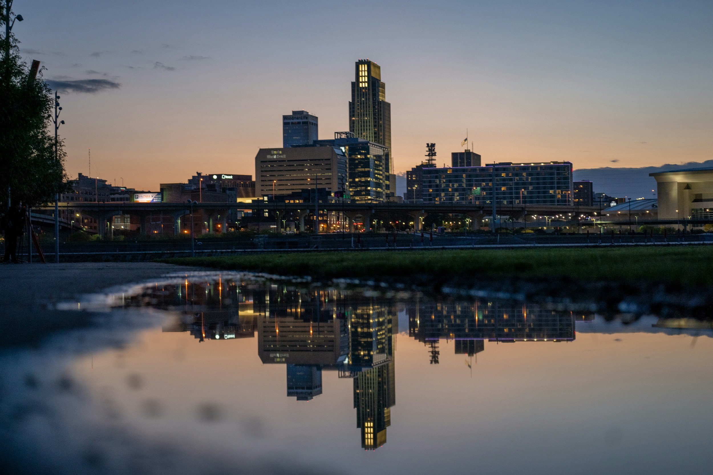 City skyline at sunset with tall buildings and a reflection in a puddle on the ground.