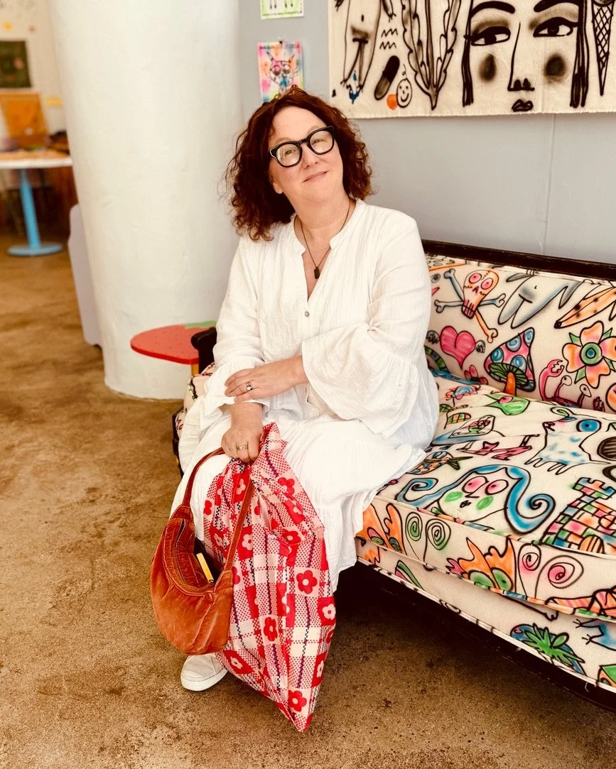 A woman with curly red hair, glasses, and a white dress sitting on a colorful, graffiti-style sofa in an art gallery or studio.