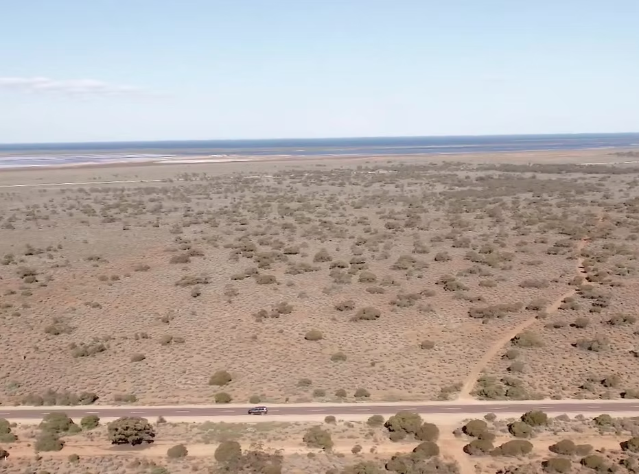 Aerial view of a desert landscape with sparse vegetation, a road running through the foreground, and a body of water in the background under a clear blue sky.