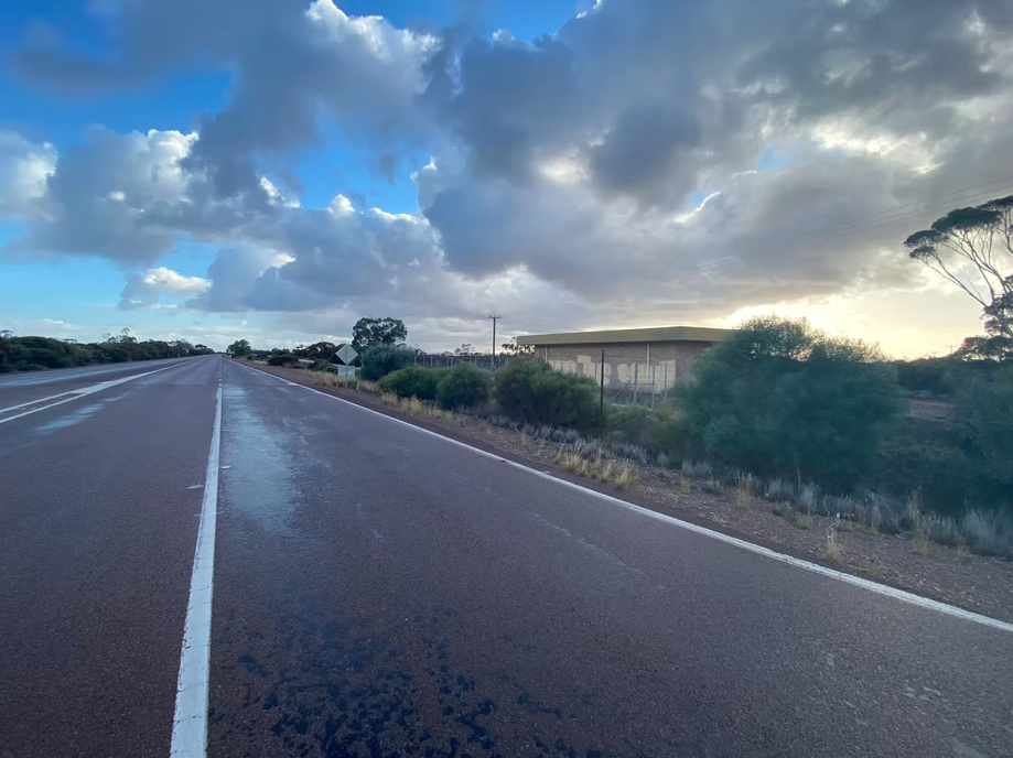 Empty highway road with a building and trees on the side, under a cloudy sky at sunset.