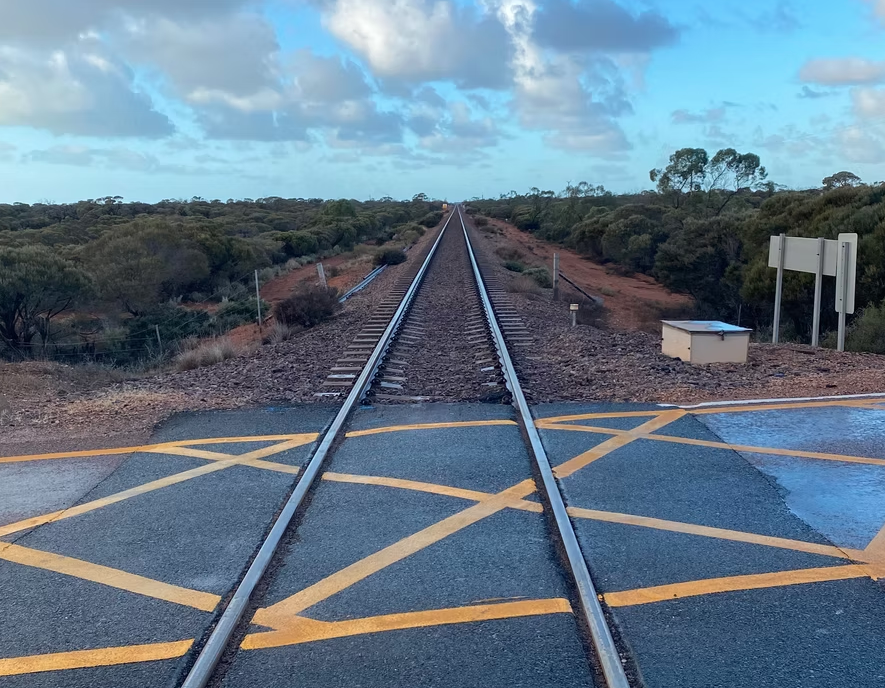 A set of railway tracks extending into the distance with safety markings on the road at a crossing, surrounded by natural landscape and cloudy sky.