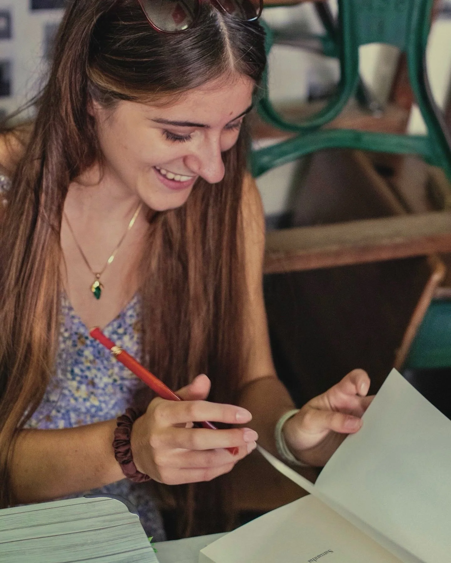A young woman with long brown hair, wearing sunglasses on her head, a patterned sleeveless top, a green pendant necklace, and a dark scrunchie on her wrist, is smiling and writing with a red pen in a notebook at a table. She is holding a piece of paper in her other hand.