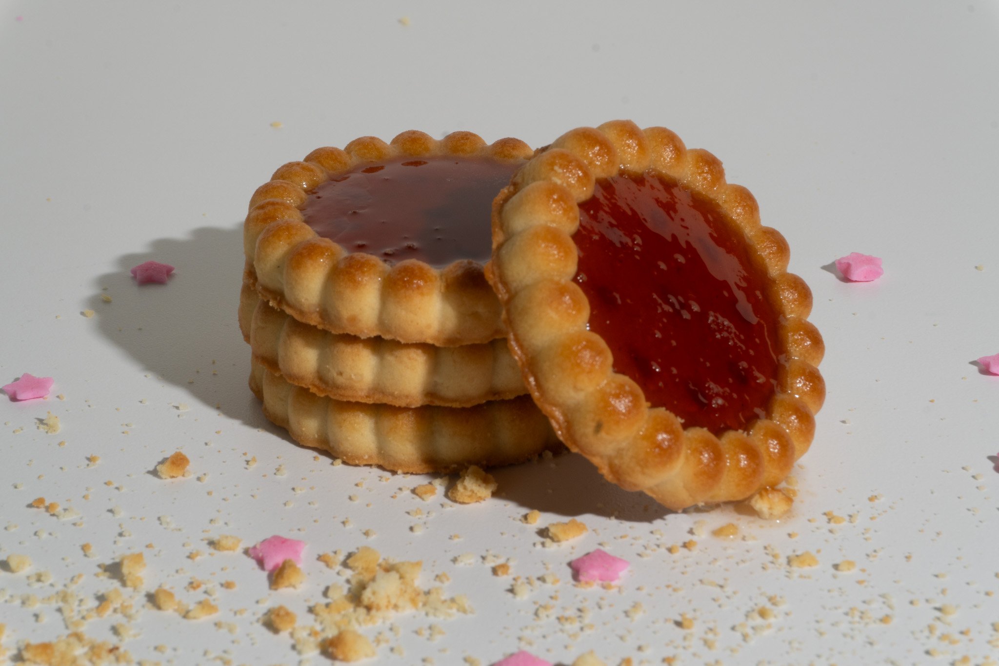 Stack of four tart cookies with a golden, ridged crust and red jam filling, placed on a white surface with scattered crumbs and pink flower-shaped confetti.