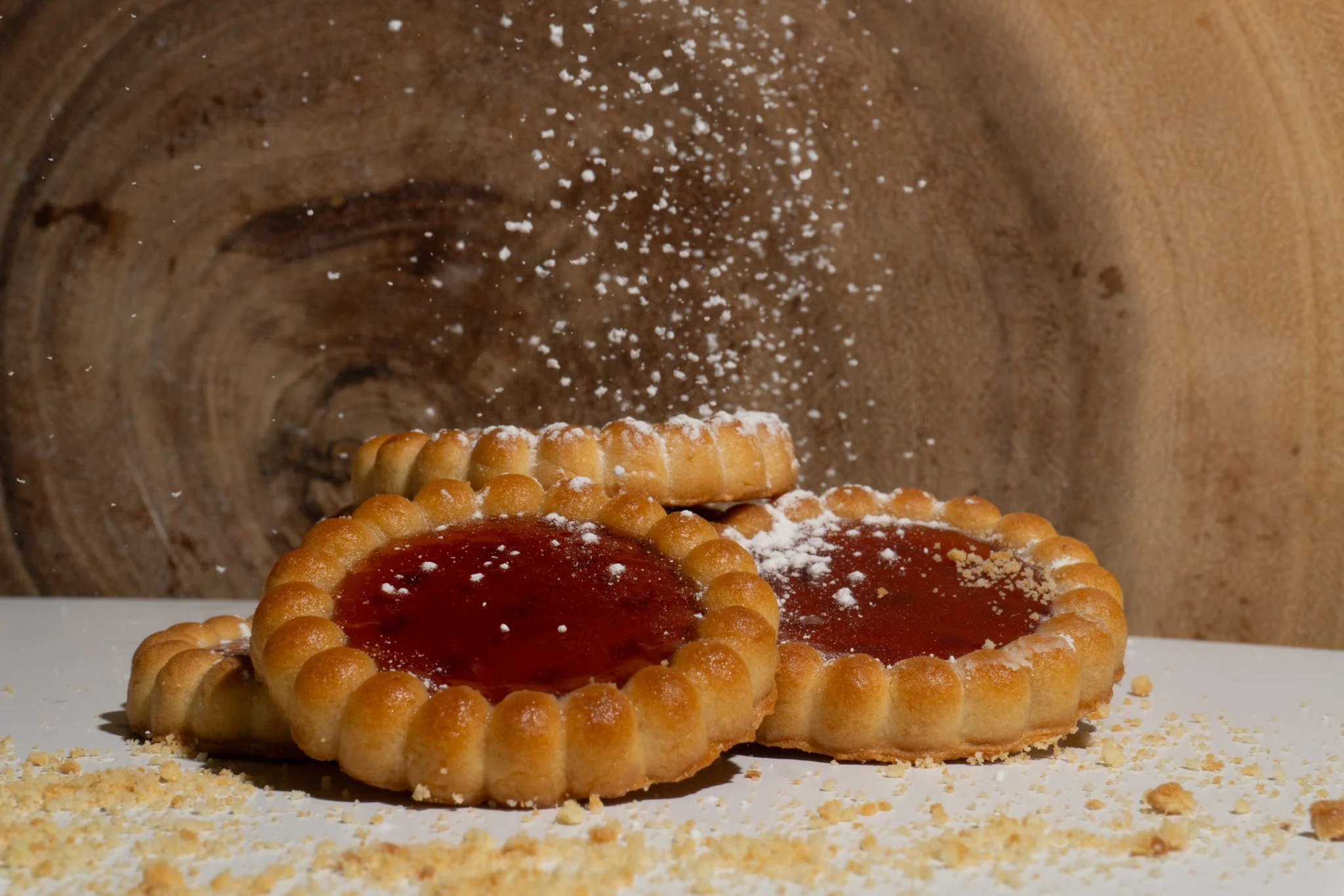 Three round cookies with jam in the center and a fluted edge, dusted with powdered sugar, on a white surface with crumbs scattered around.