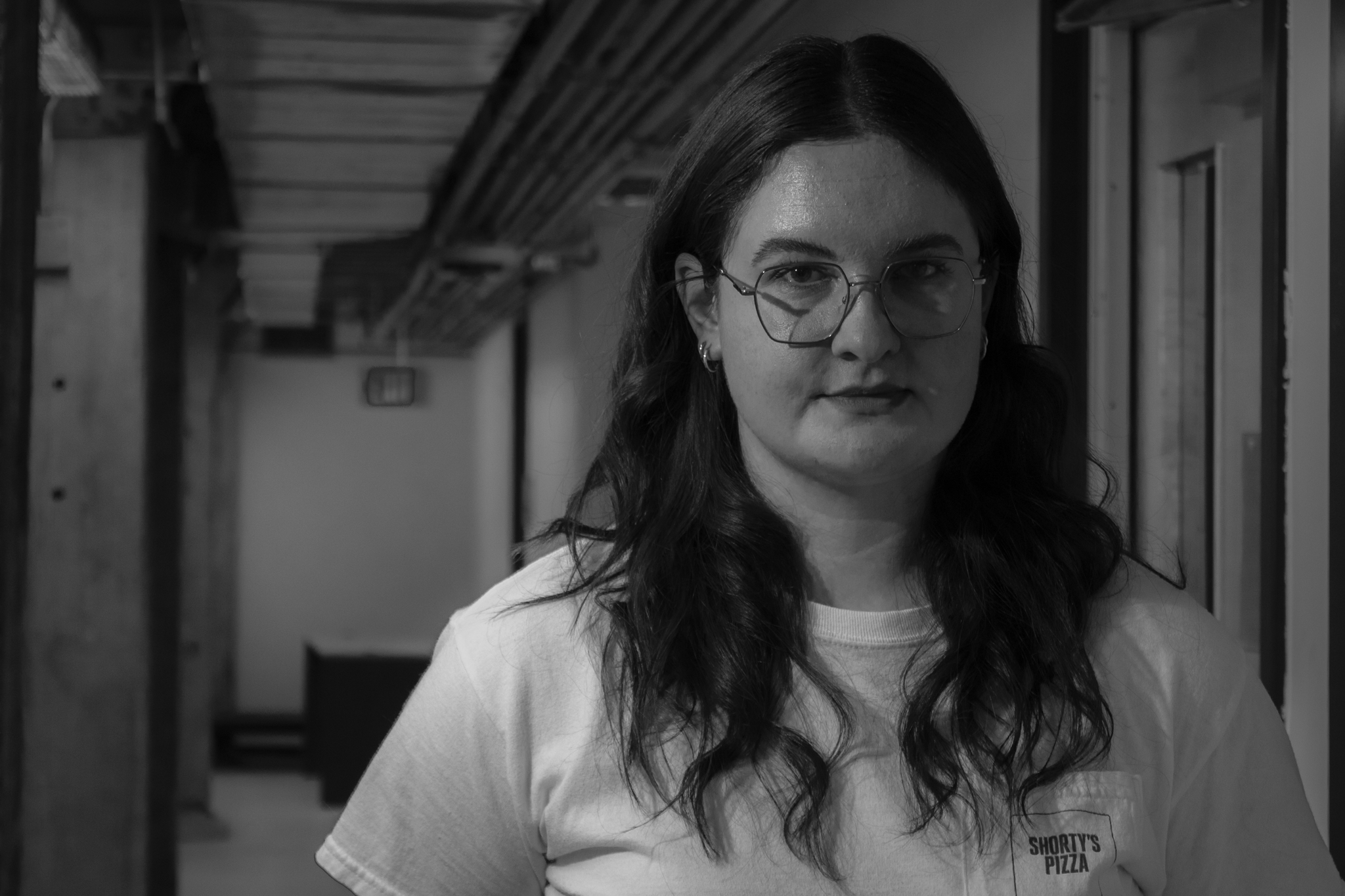 A woman with long dark hair, glasses, and earrings, standing in a hallway with a serious expression. She is wearing a short-sleeved white t-shirt with a logo that says "Shorty's Pizza." The hallway has wood and metal structures, and is dimly lit.