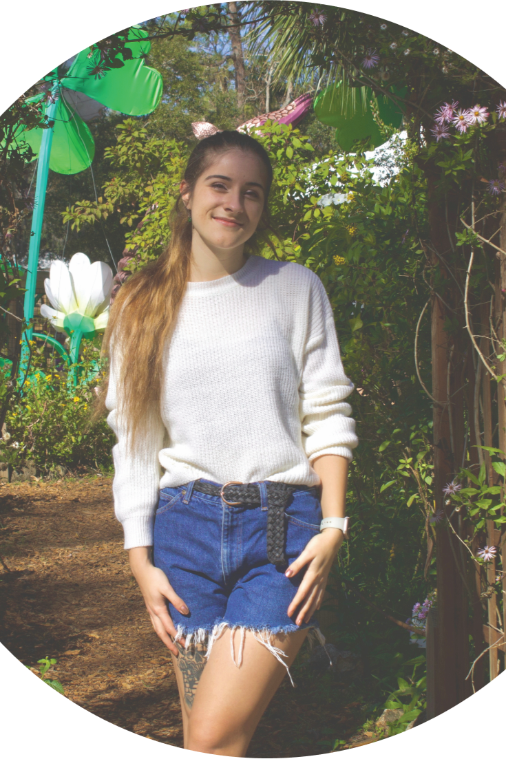 A young woman with long hair in a white sweater and denim shorts standing in a lush garden with colorful plants and large decorative flowers.