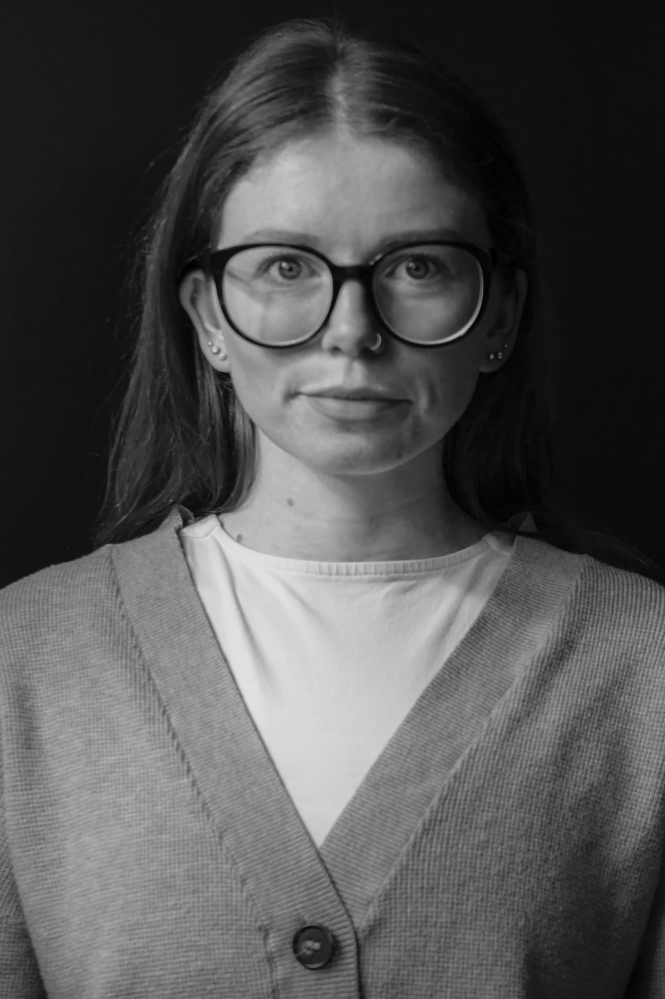 Black and white portrait of a woman with glasses, long hair, and multiple earrings, wearing a buttoned cardigan over a t-shirt, against a dark background.