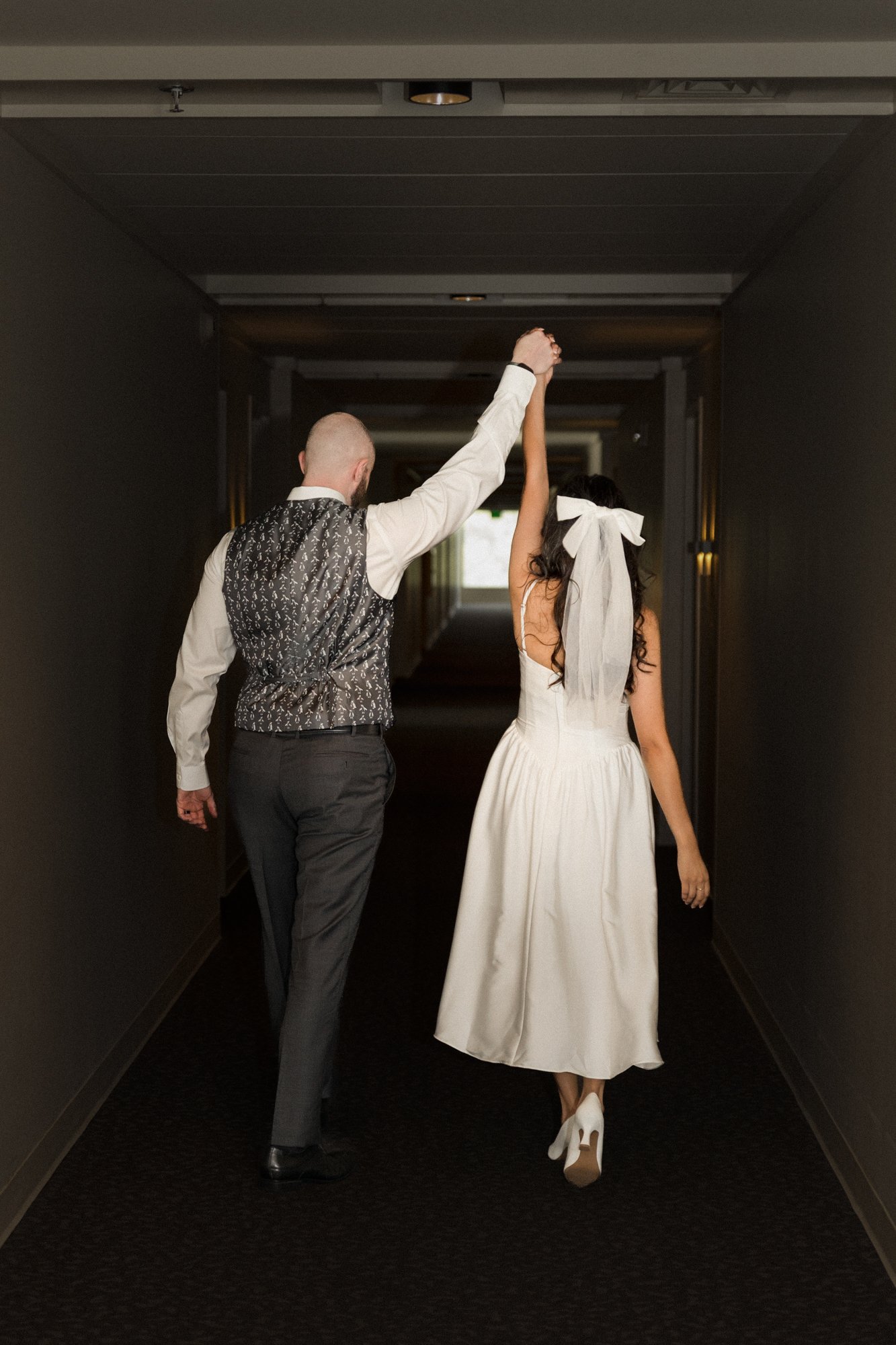 A couple holding hands while walking down a hallway, with the woman wearing a white dress and heels, and the man dressed in a vest and trousers.