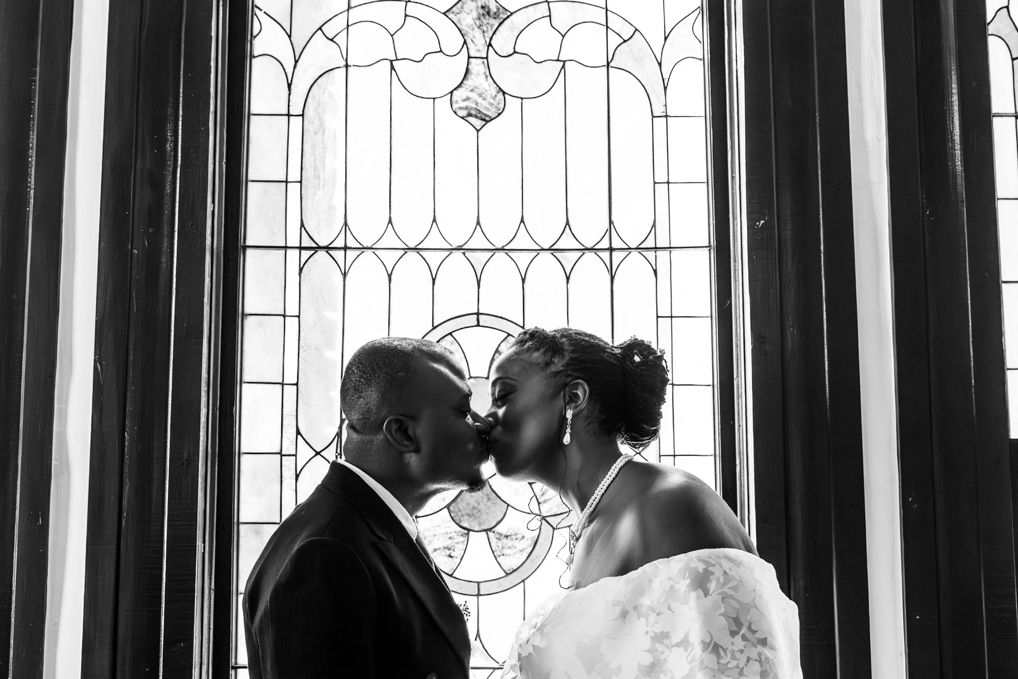 A black-and-white photo of a couple kissing in front of a stained glass window. The man is wearing a suit and the woman is in a wedding dress with earrings, a necklace, and an updo hairstyle.