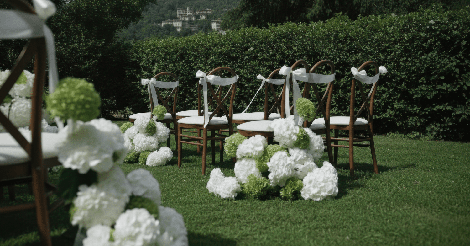 Wedding ceremony setup with wooden chairs decorated with white ribbons, surrounded by white and green floral arrangements on a grassy outdoor area.