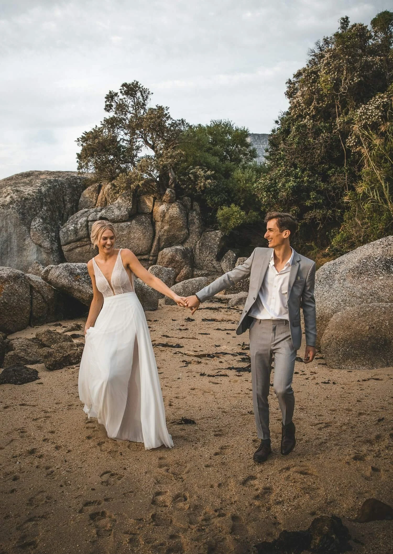 A couple in formal attire holding hands on a sandy beach with large rocks and green trees in the background, smiling at each other.