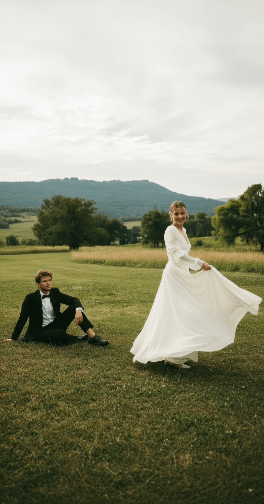 A bride in a white wedding dress stands on a grassy field, smiling, while a groom in a black tuxedo sits on the grass nearby, set against a backdrop of trees and mountains.