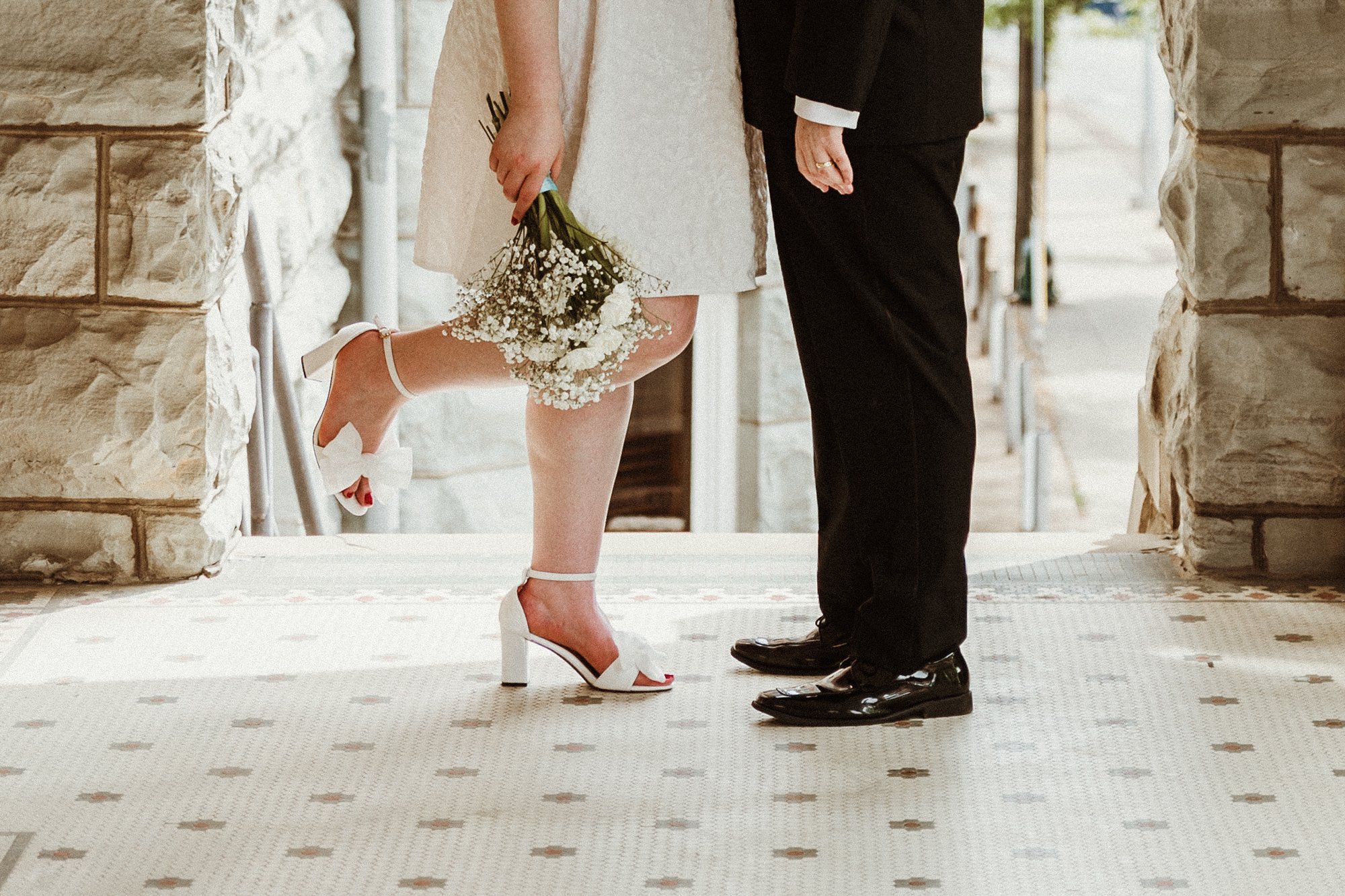 A bride and groom standing close together during their wedding ceremony, with only the lower halves of their bodies visible. The bride is wearing a white dress, white high-heeled shoes, and holding a bouquet of white flowers. The groom is dressed in a black suit with shiny black shoes. They are standing on a patterned rug between stone columns.