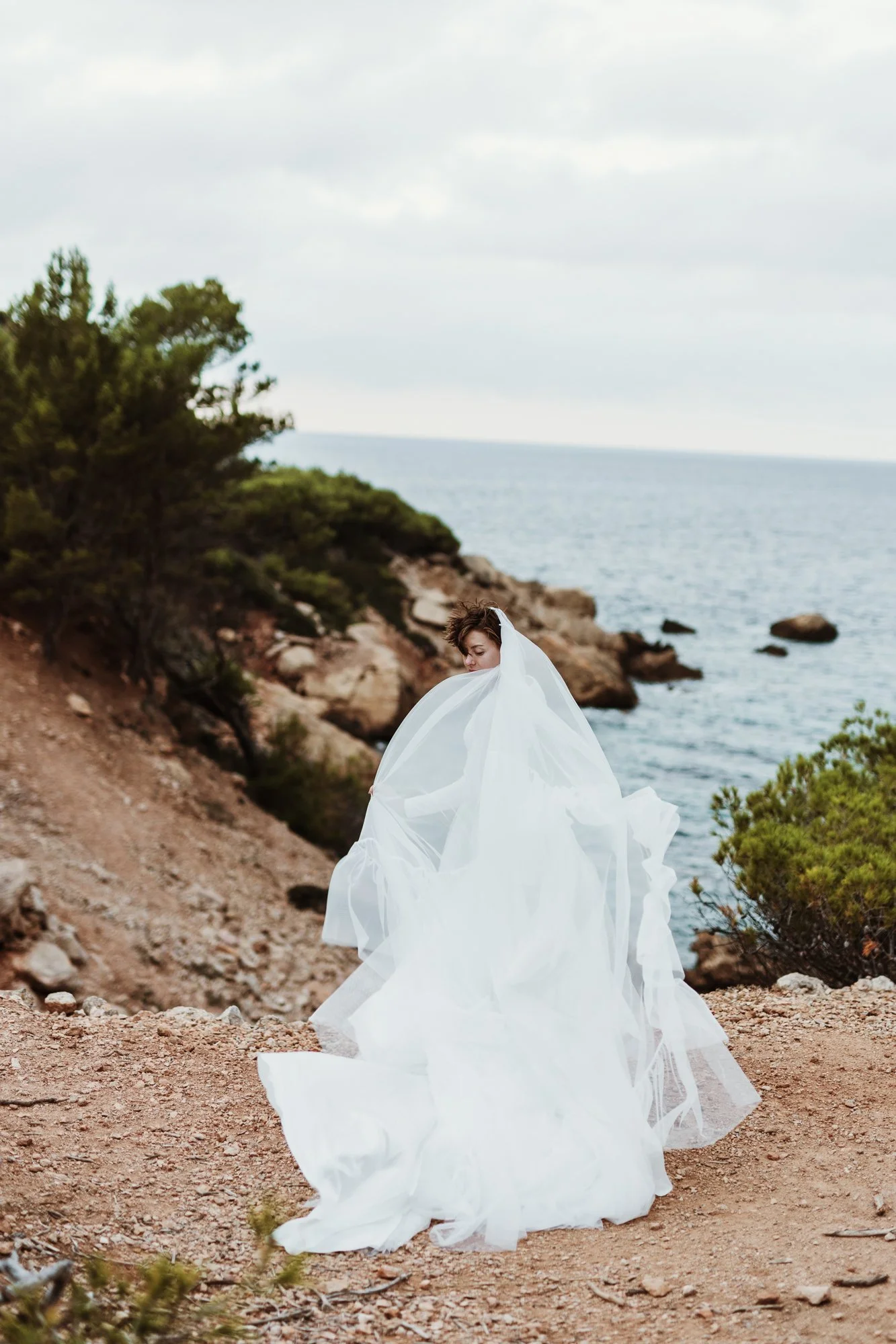 A person in a bridal gown is standing on a rocky coastal landscape with the ocean and cloudy sky in the background.