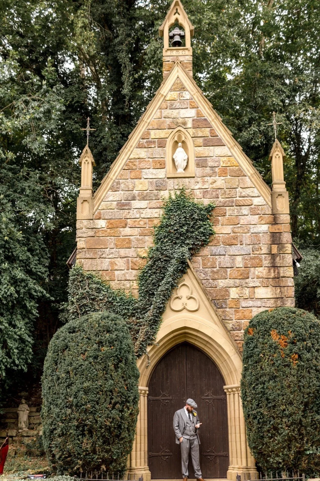 A man in a gray suit and gray flat cap standing in front of a large wooden church door, holding a drink and looking at his phone. The church is made of brick and stone with ivy and greenery around it, and has Gothic architectural features.