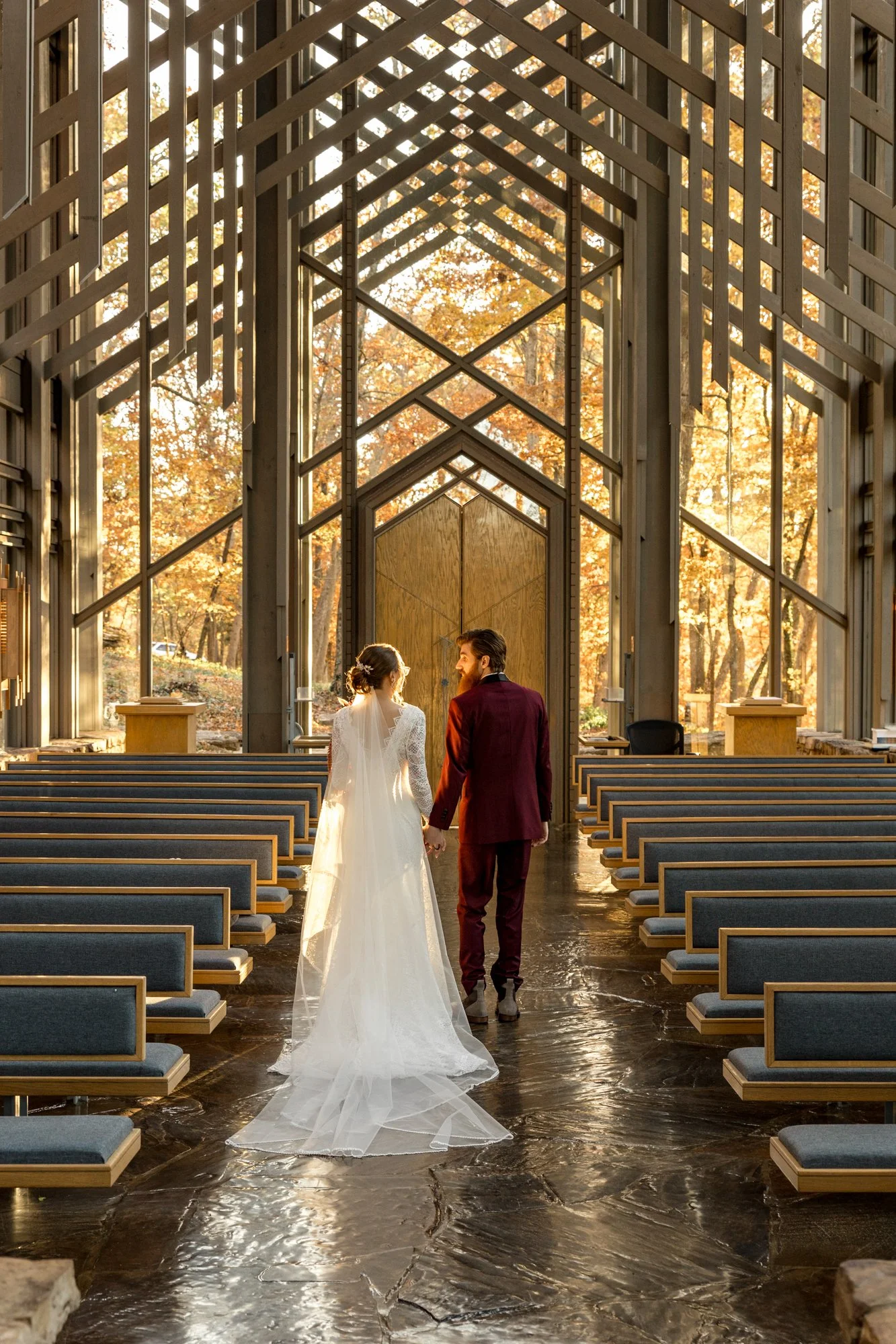 A bride and groom holding hands inside a modern church with large glass windows and wooden architectural details, during autumn with trees visible outside.