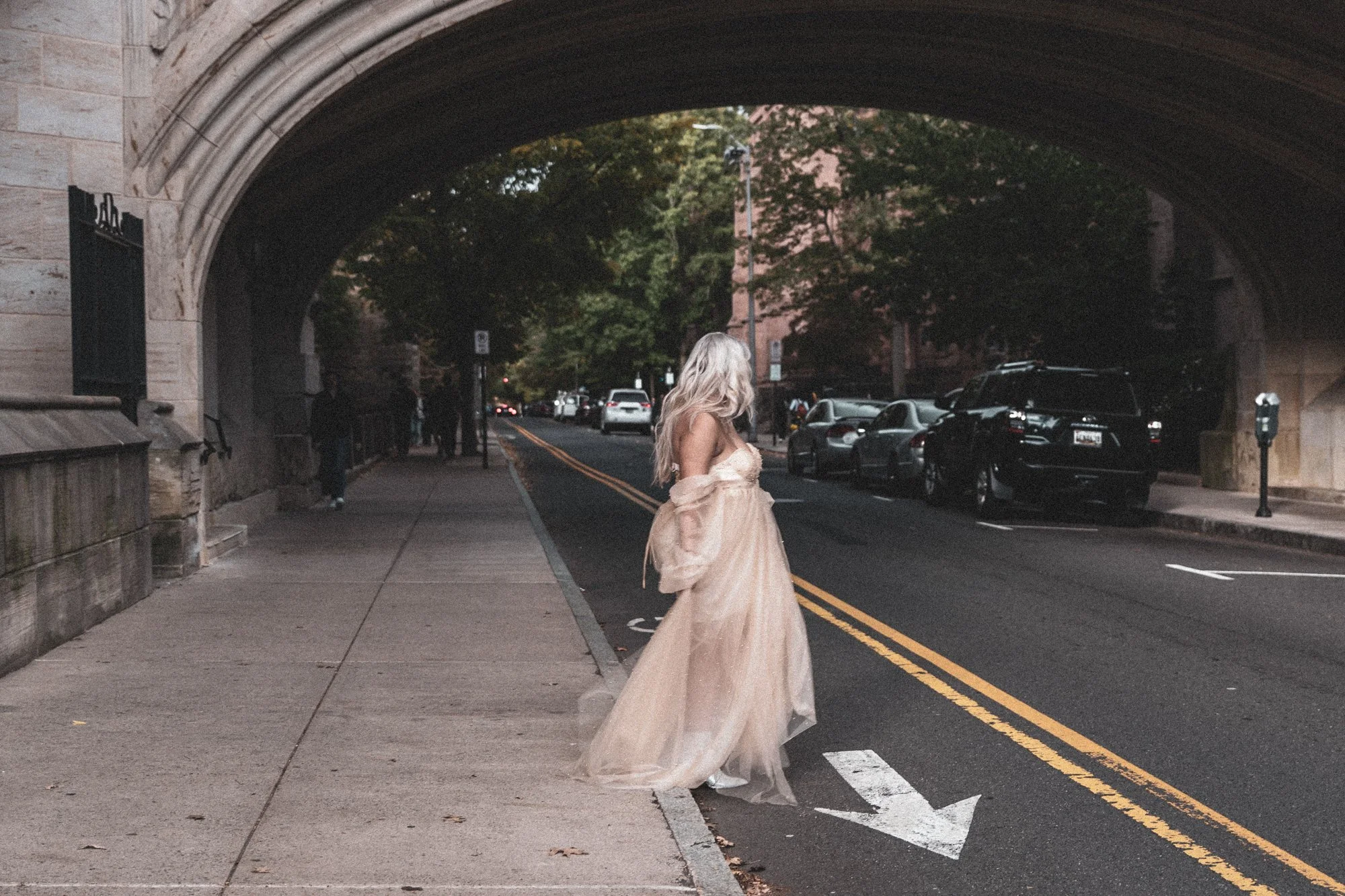 A woman with long blonde hair wearing a flowing, beige dress standing on a city sidewalk under a stone arch bridge. Cars are parked along the street on the right side, and trees and buildings are visible in the background.