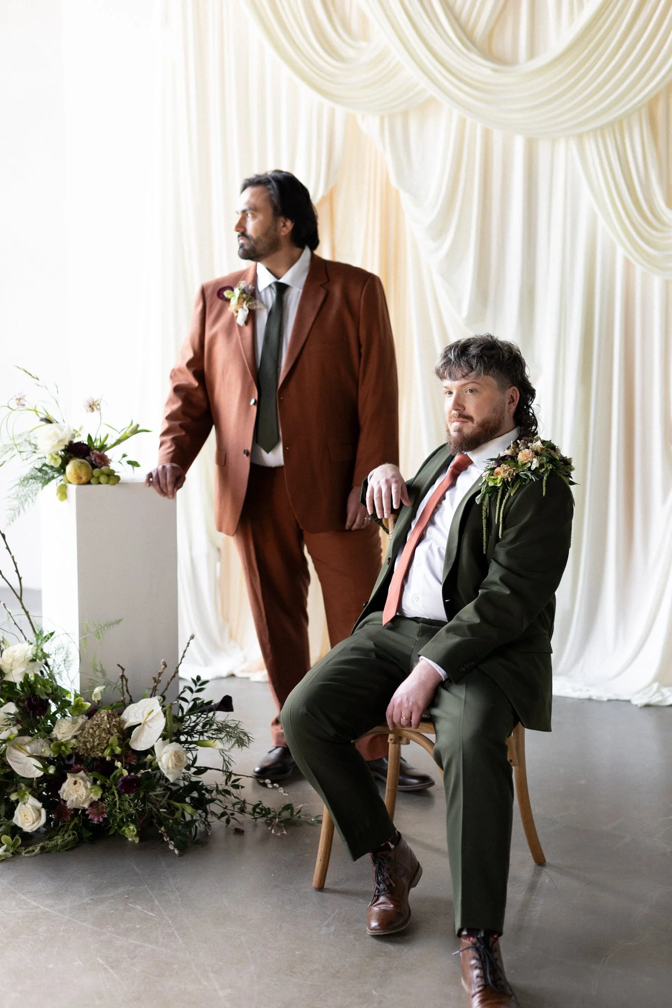 Two men in suits with floral boutonnieres at a wedding ceremony; one man is standing and the other is seated, both with serious expressions; floral arrangements and draped fabric decor adorn the background.