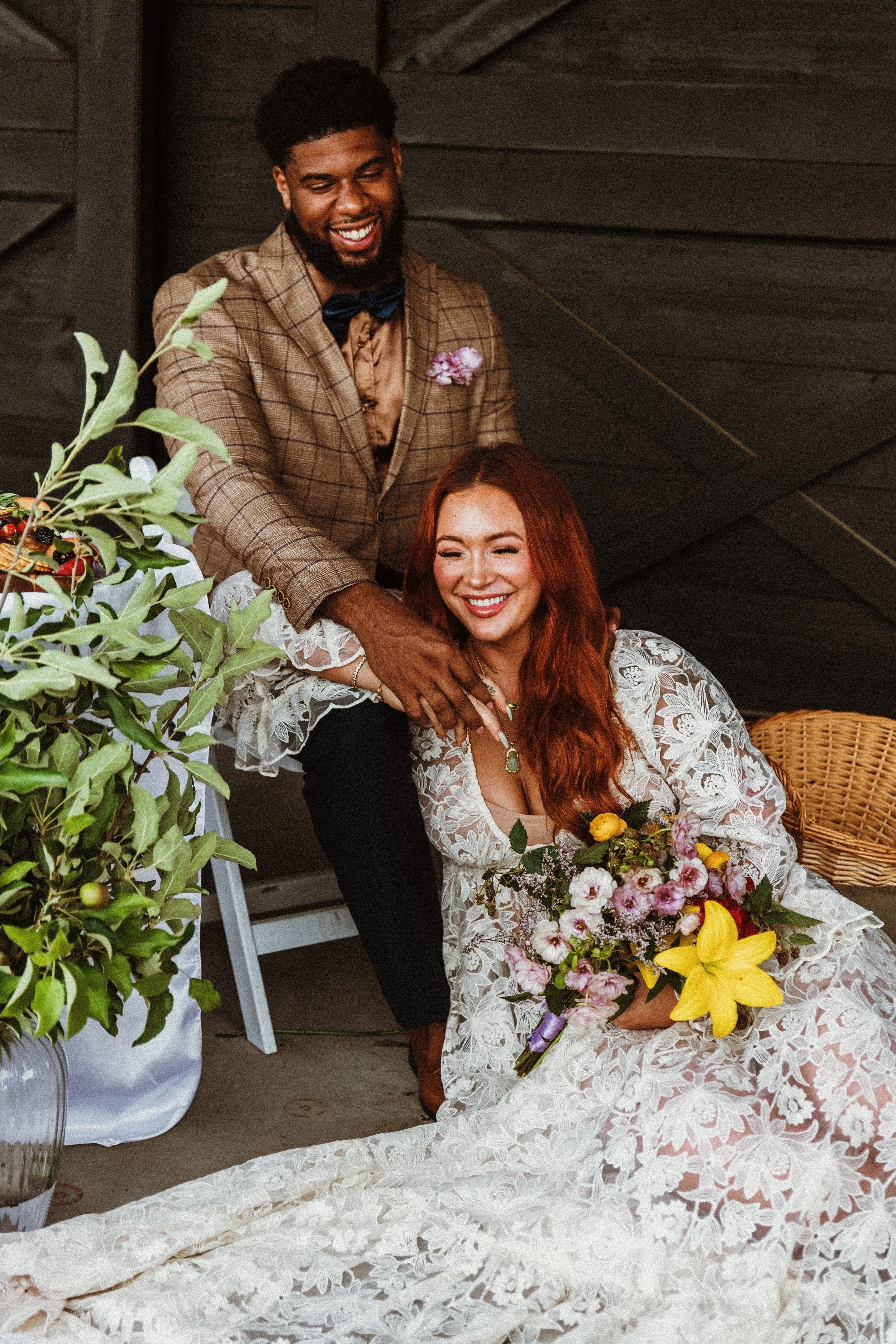 A man in a checked blazer and bow tie stands behind a woman in a white lace dress, both smiling, with the woman holding a large floral bouquet. They are indoors with dark wooden walls and some green plants nearby.