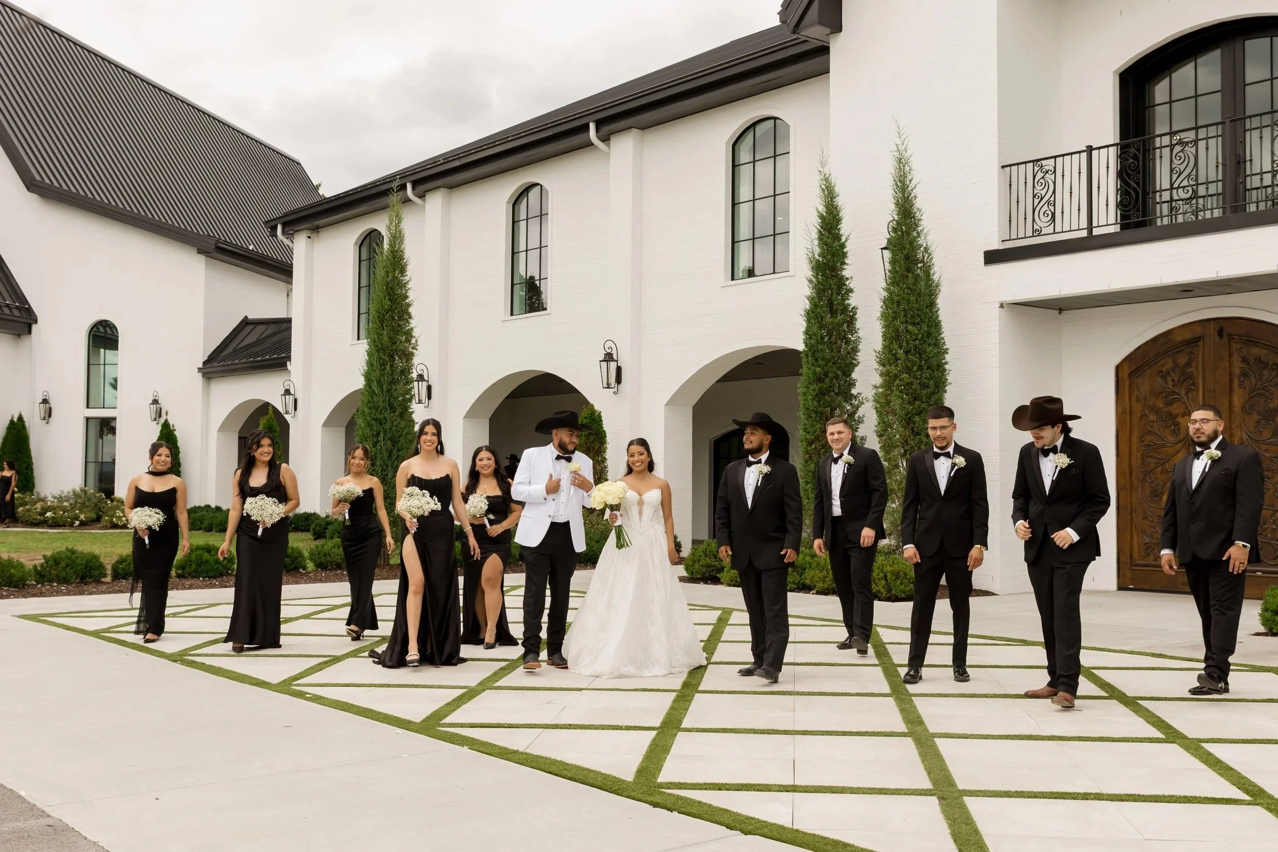 A group of bridesmaids and groomsmen standing outside a large white building with arched windows and tall trees, dressed in formal wedding attire, holding flowers and smiling.