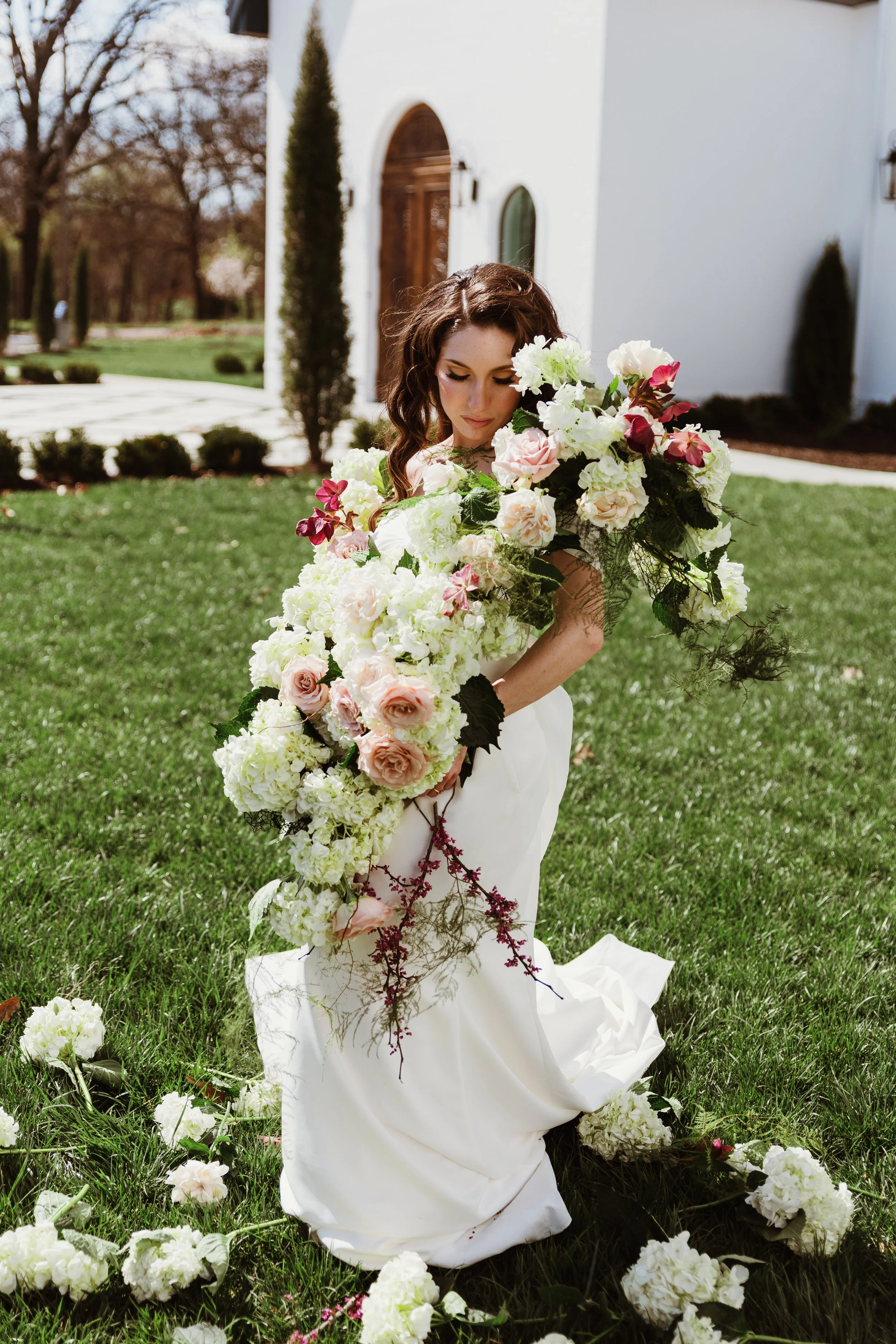 A woman wearing a white dress is kneeling on green grass outside a white building, holding a large bouquet of white and blush pink flowers.