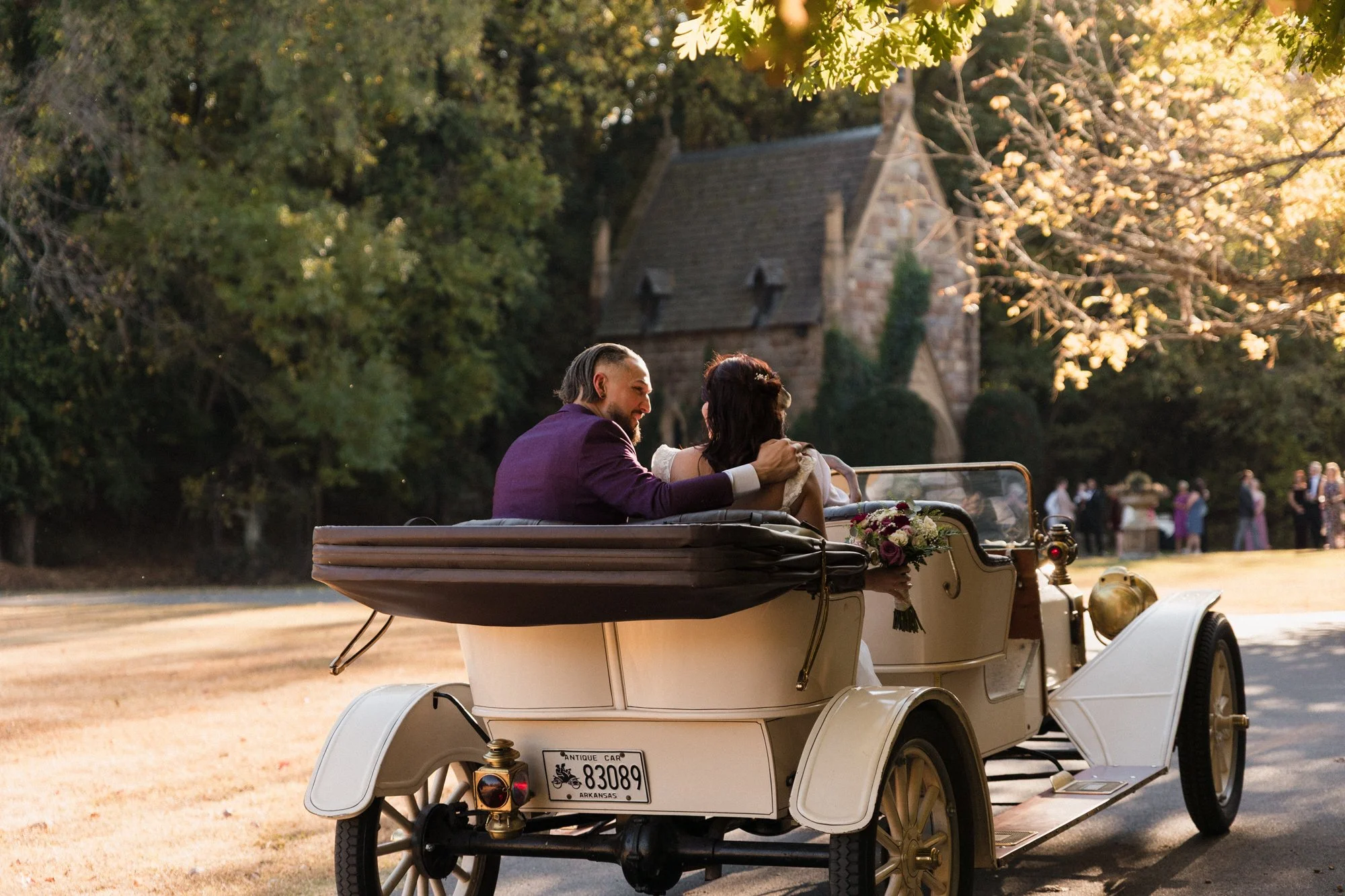 A couple dressed in wedding attire riding in a vintage golf cart decorated with flowers through a park with trees and a stone building in the background during sunset.
