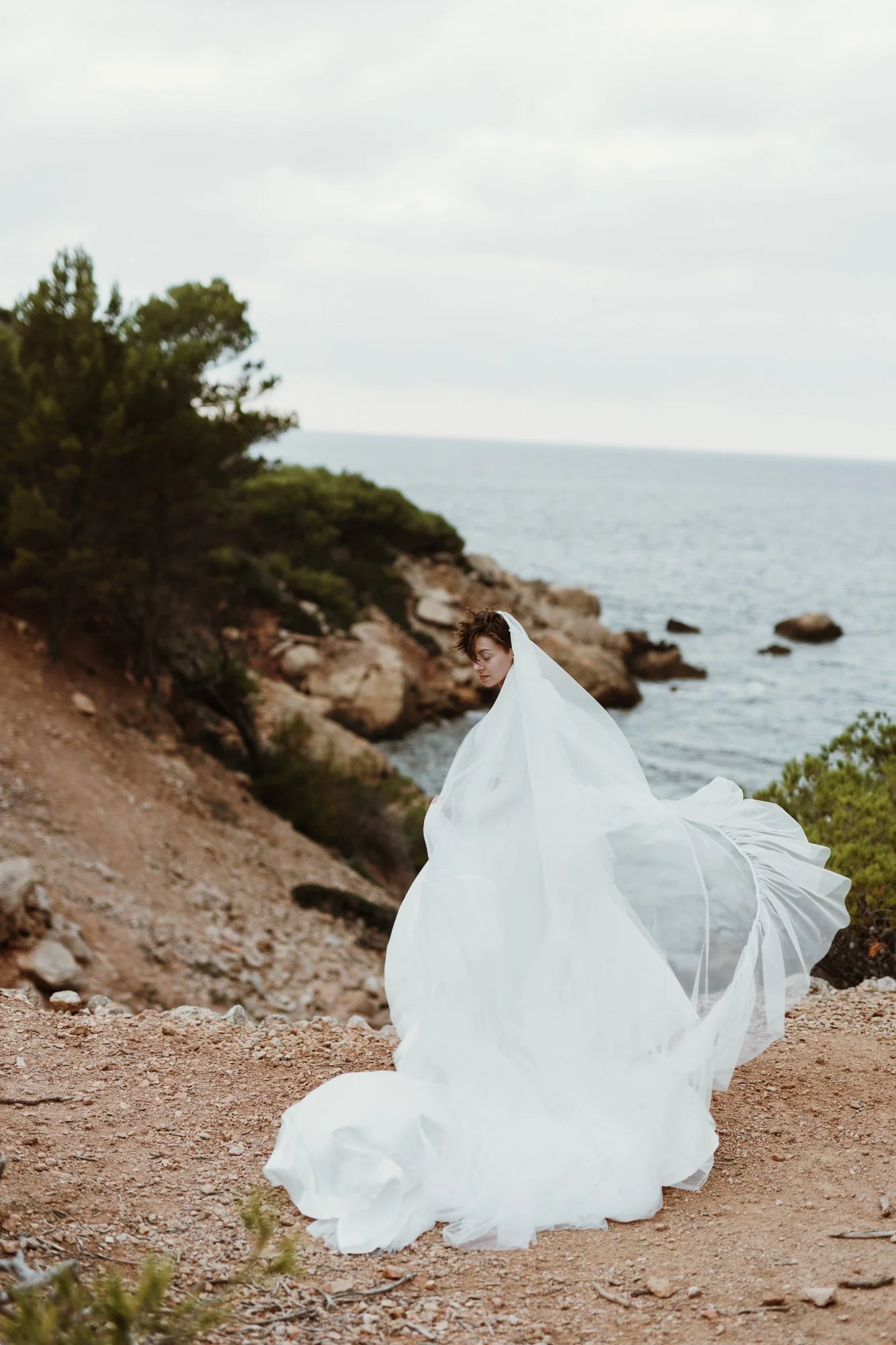A woman in a flowing white dress stands on a rocky, dirt path by the coast with the ocean and cloudy sky in the background.