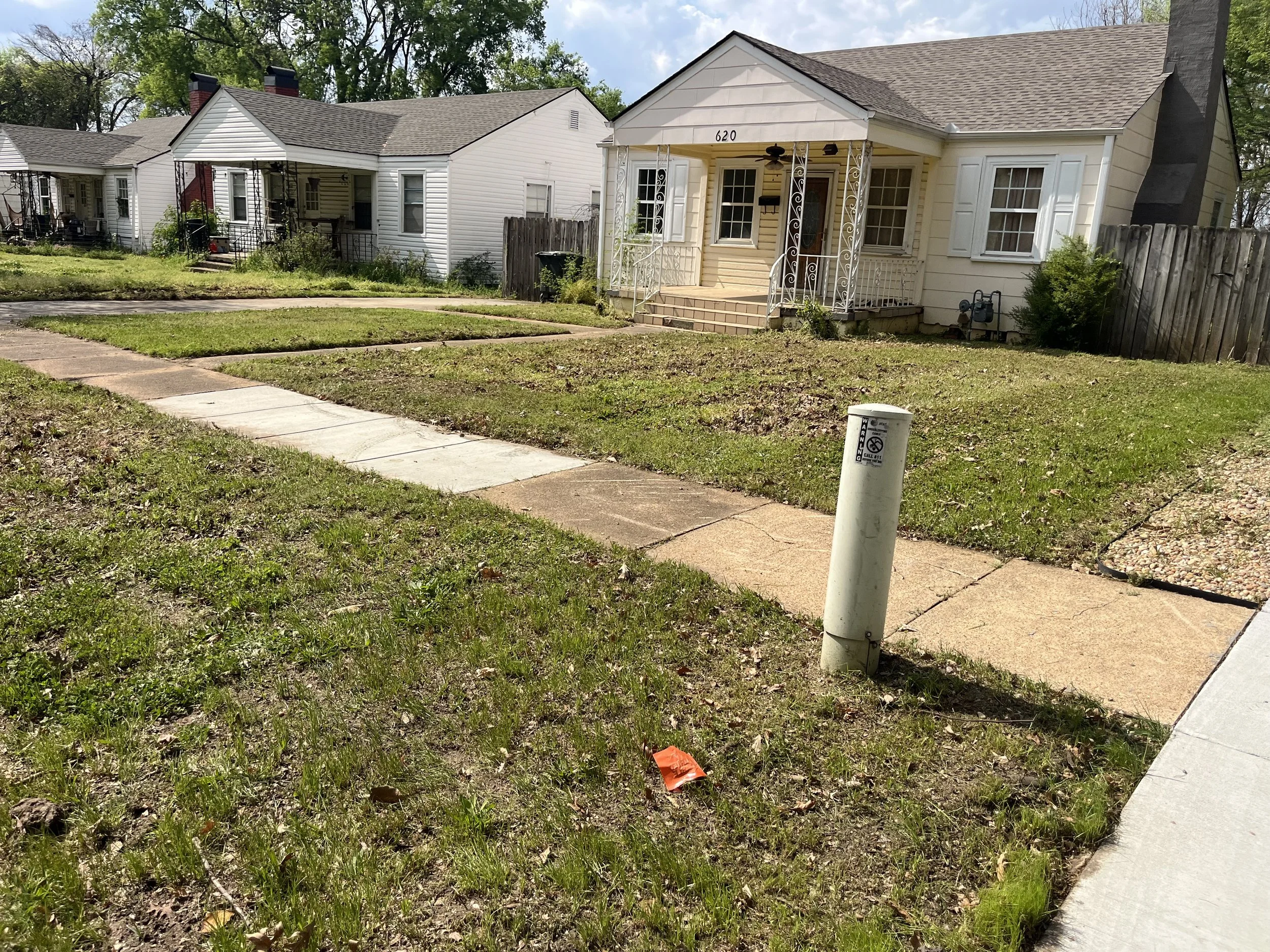 A suburban neighborhood with a grassy lawn, concrete sidewalks, and two single-story houses with porches. One house is cream-colored with white shutters and a small front porch. The other house is white with darker trim. There is a utility pole in the grass near the sidewalk and some trees in the background.