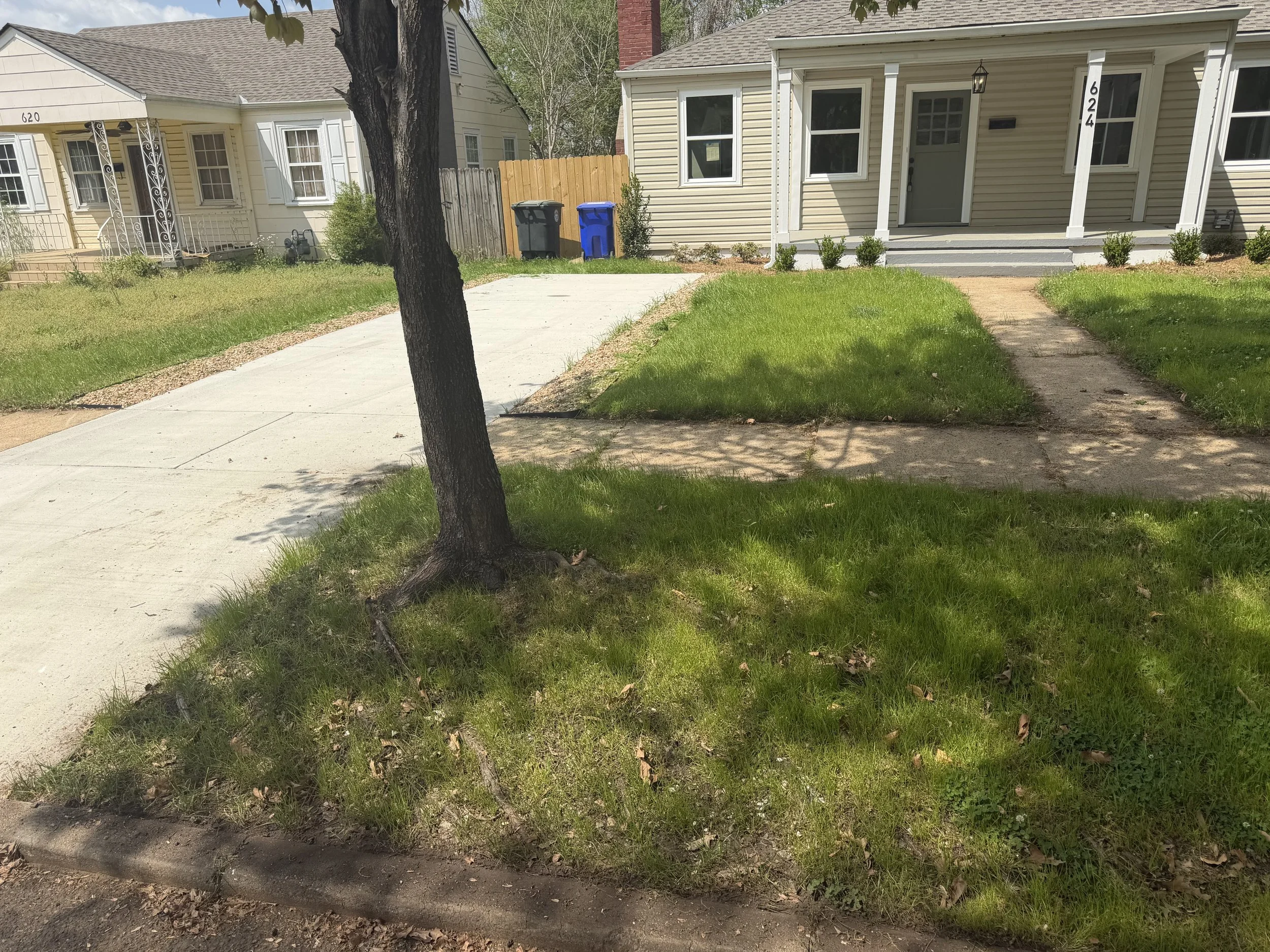 A suburban front yard with a concrete sidewalk, a grassy lawn, and a house with white siding, front porch, and small bushes. There is a tree in the foreground and two trash bins near the fence.