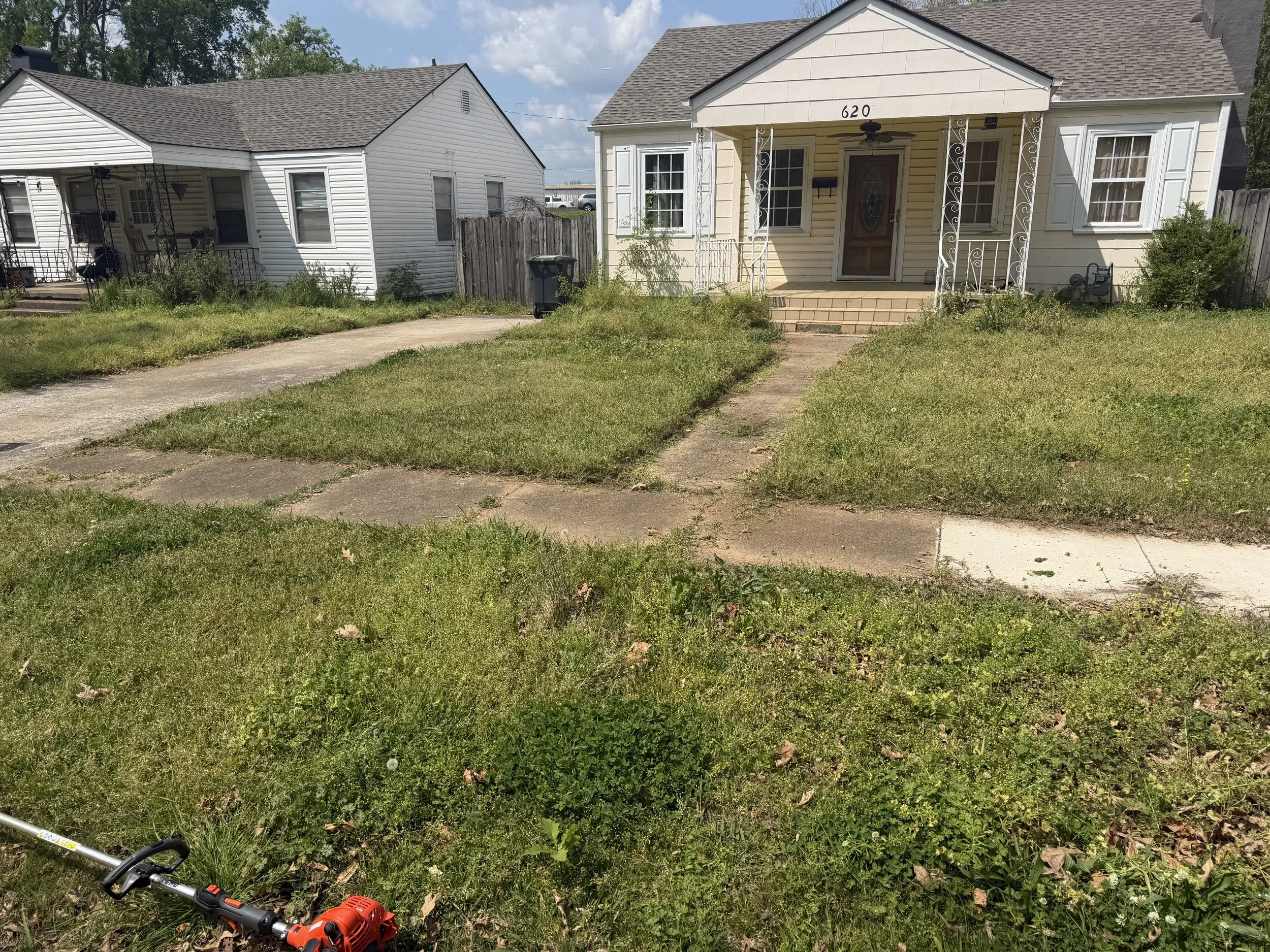 Front yard of a house with a concrete walkway, grass, and uneven patches of weeds. The house has a small porch with decorative iron supports, a brown door, and white siding. There is a neighboring house to the left with similar siding and a porch. A weed trimmer lies on the grass in the foreground.