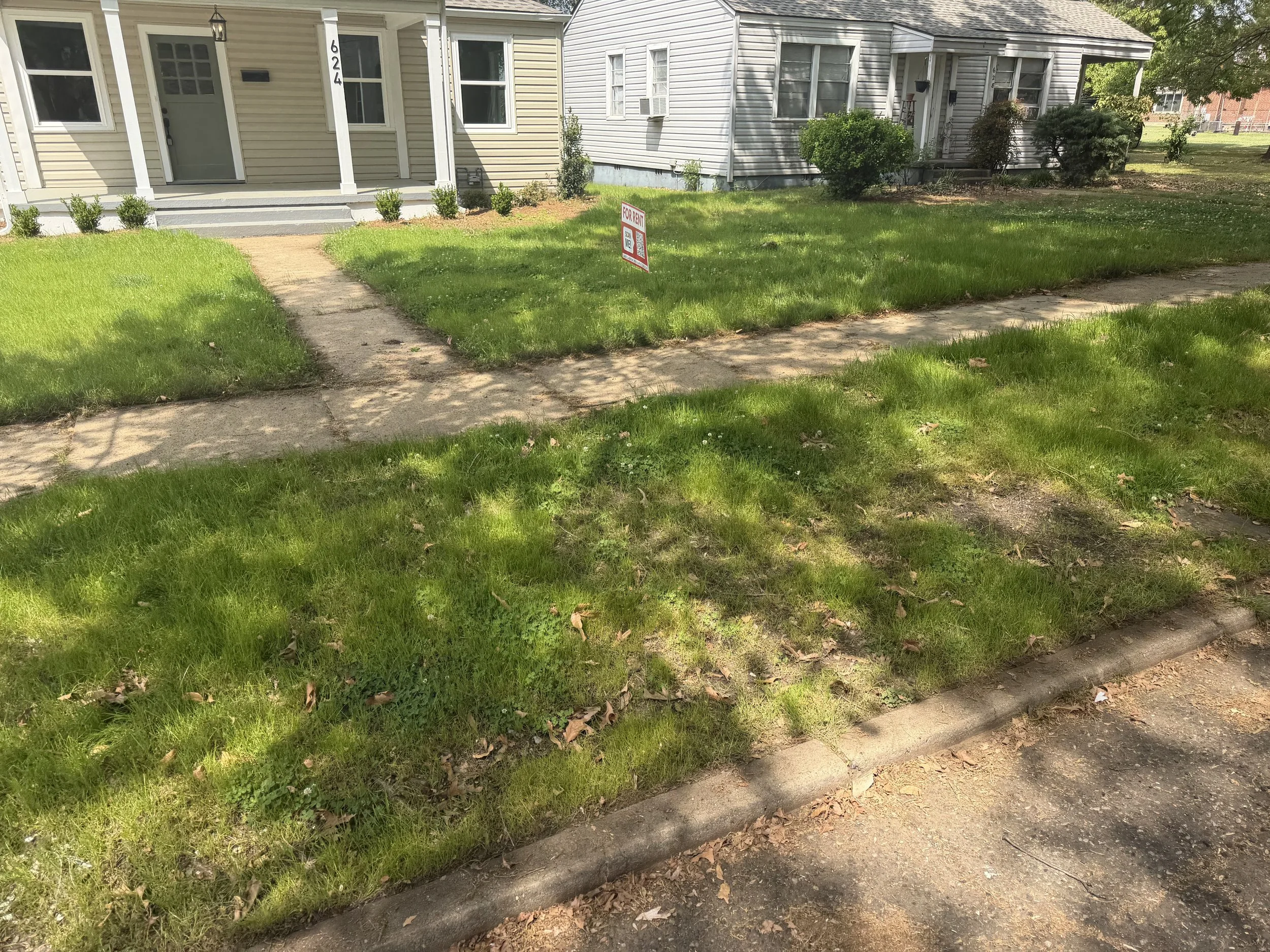 A residential front yard with a sidewalk, grass, and a house with beige siding. There is a red and white 'For Rent' sign in the yard and a house with gray siding in the background.