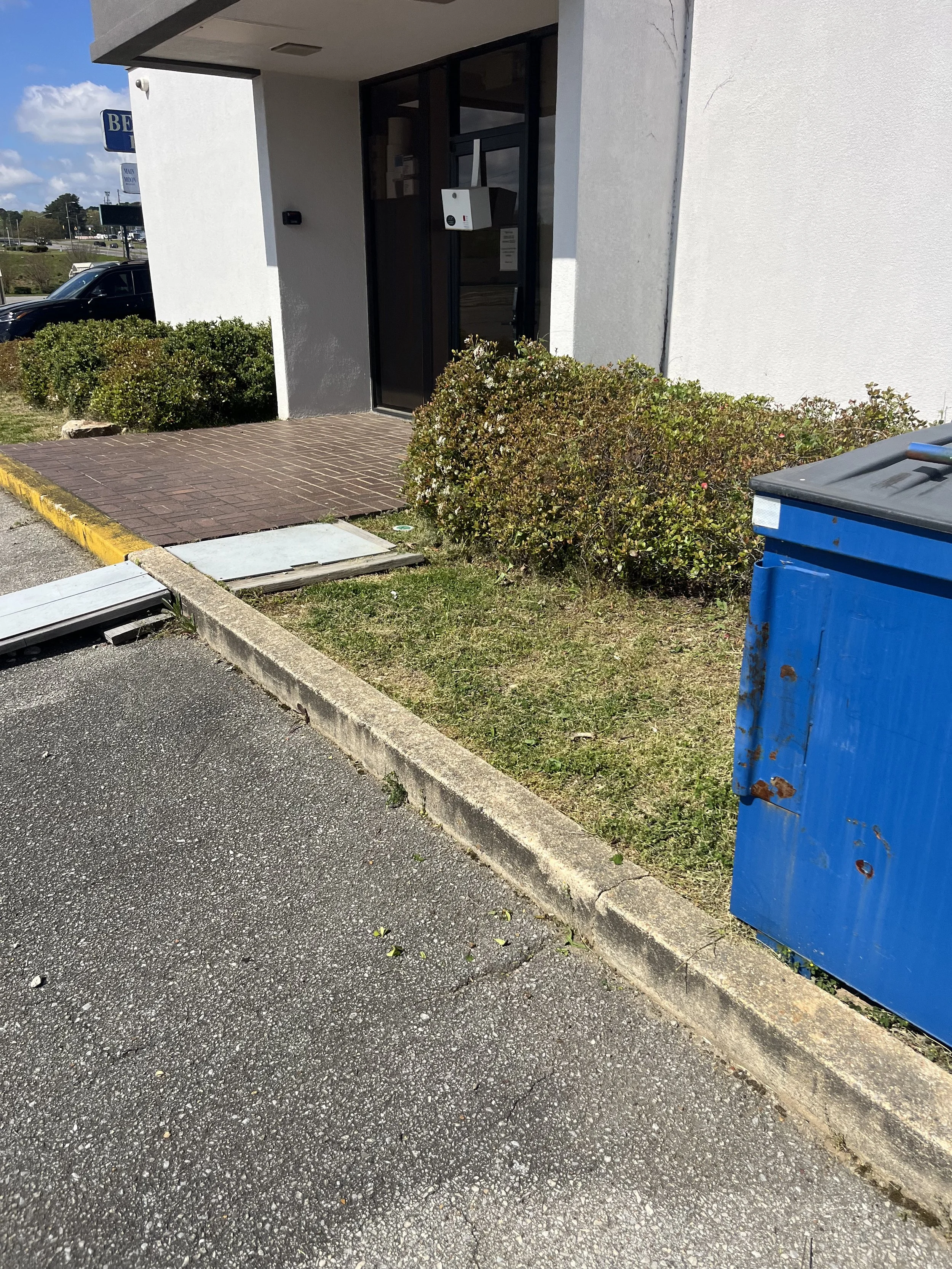 Building entrance with black door, surrounded by bushes, next to a blue dumpster, with a paved sidewalk and parking lot.