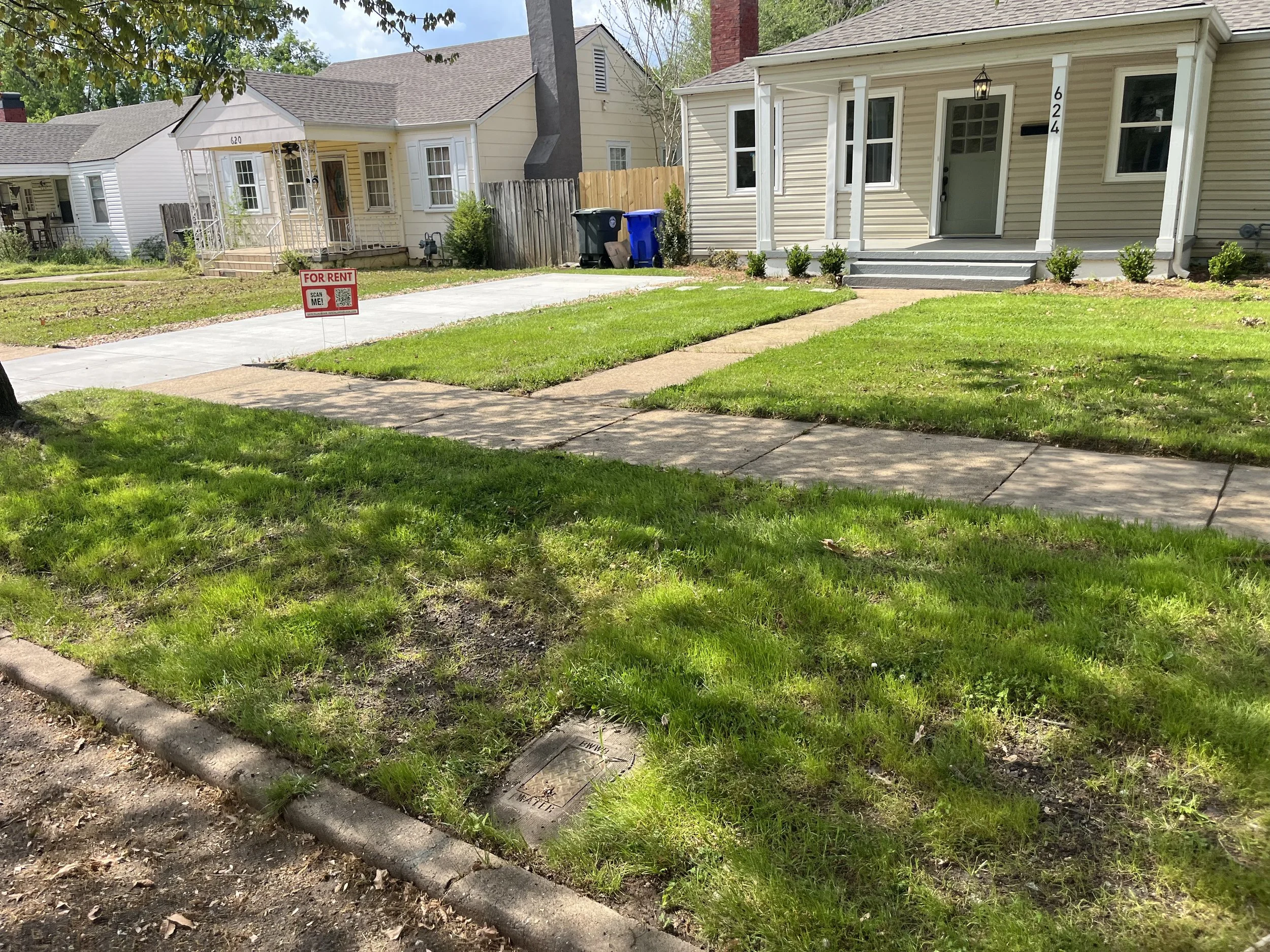 A row of houses with front yards, a sidewalk, and a for rent sign in the yard of the house on the left. The house on the right has the number 624 and a small porch with steps and a lamp. There are trees and blue recycling bins in the background.