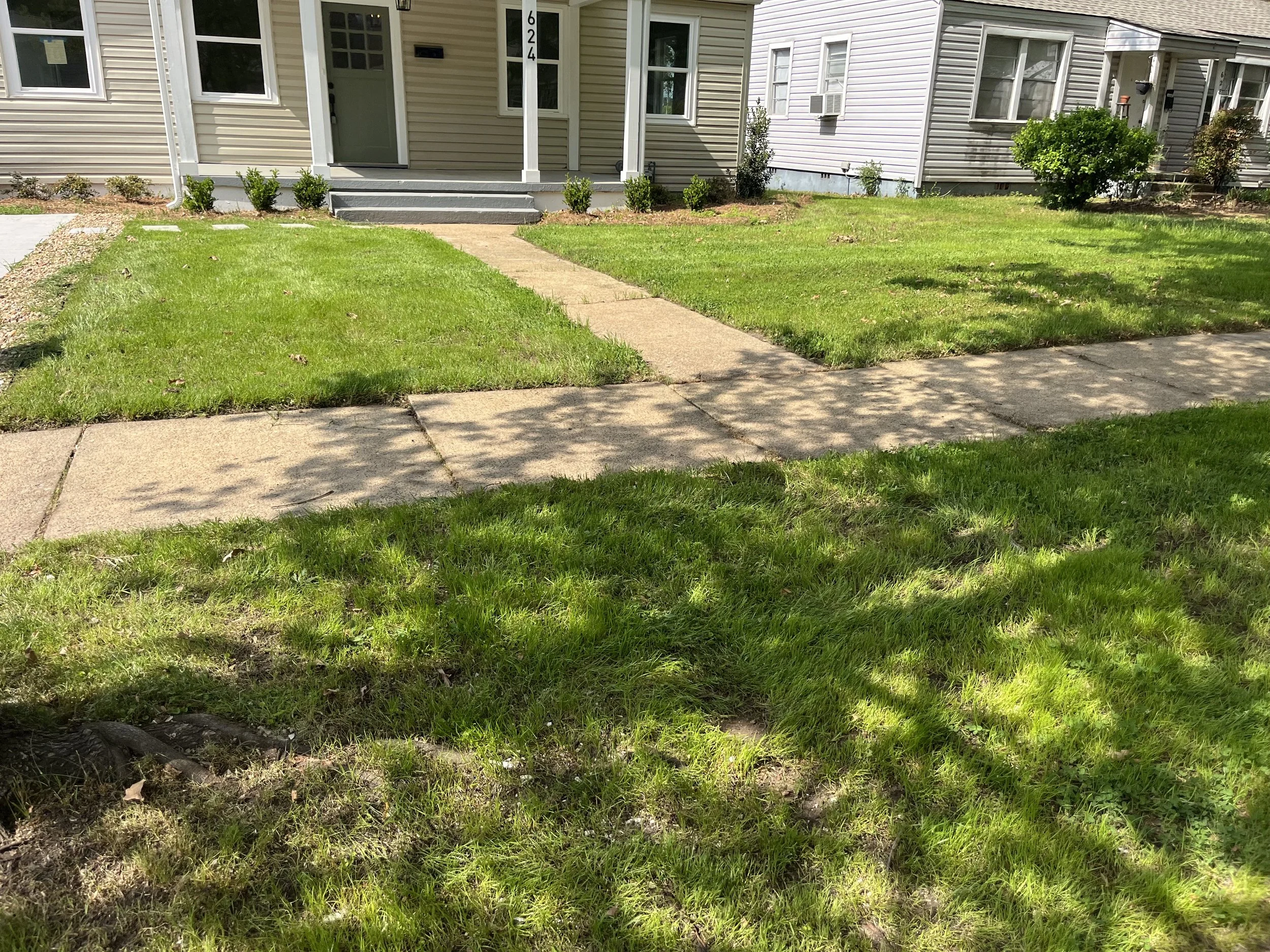 A residential front yard with a concrete walkway, green grass, small shrubs, and a house with beige siding and a porch.