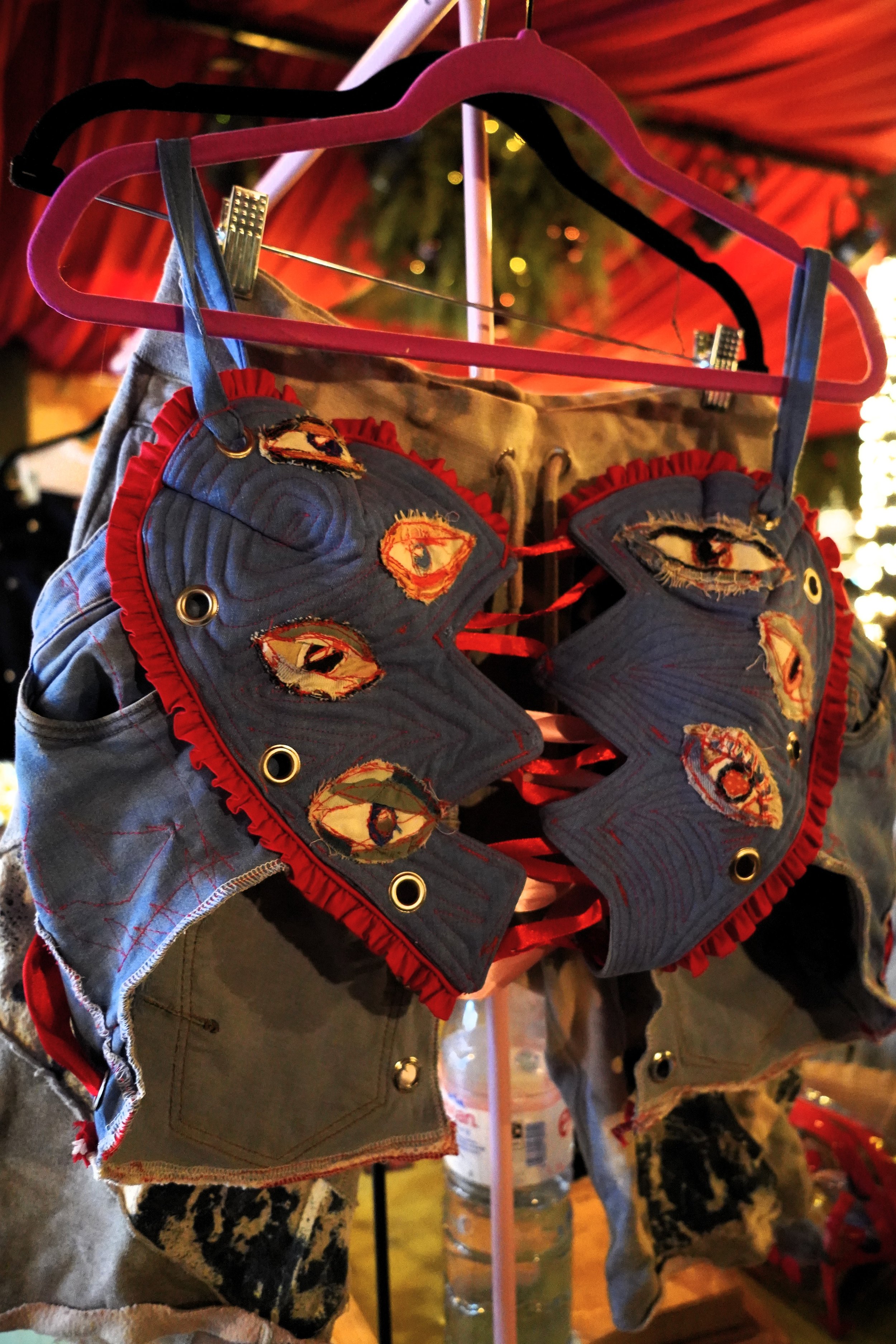A pair of denim and patterned fabric shorts with eye-shaped fabric patches and red trim, displayed on pink and black hangers.