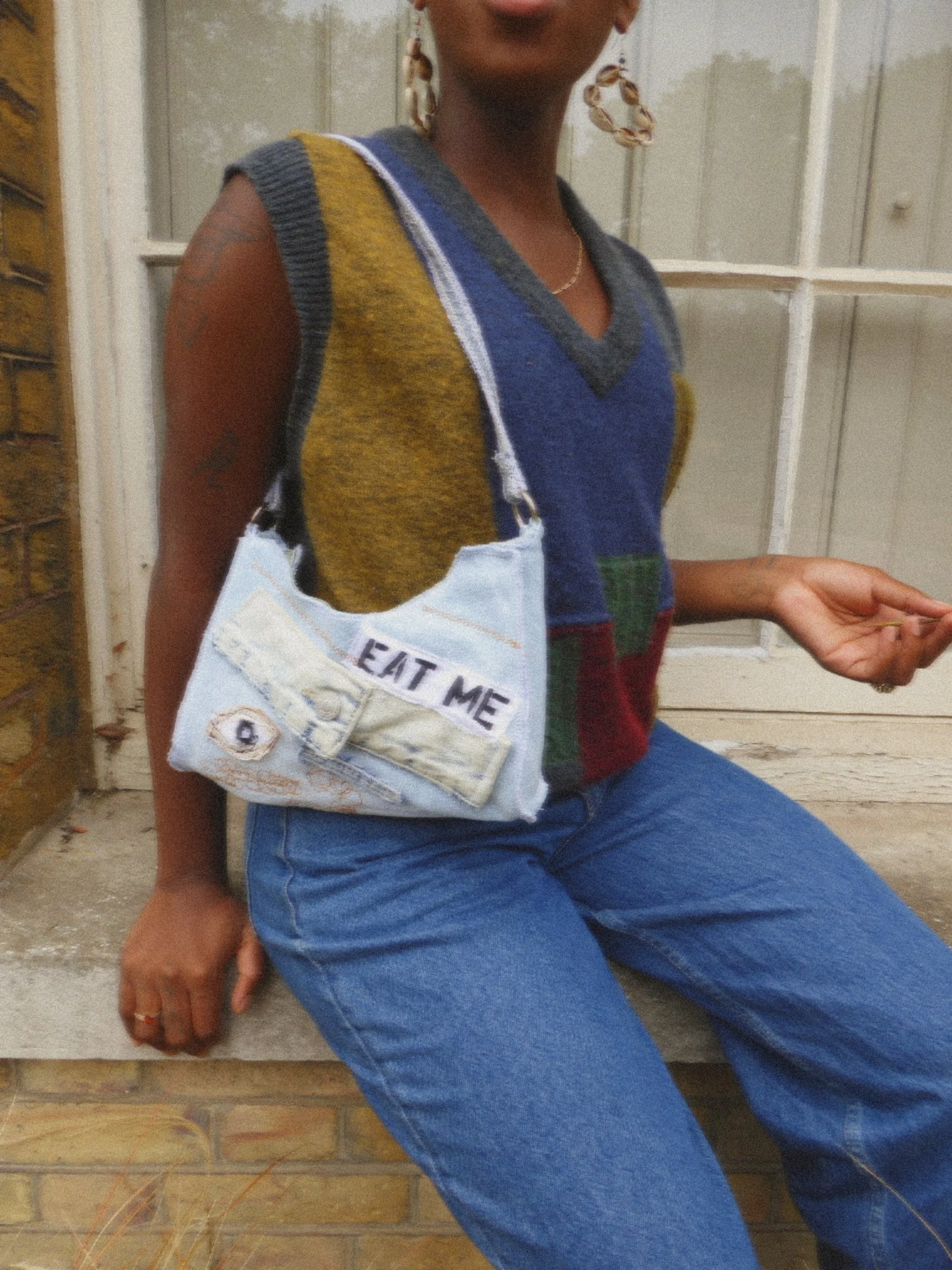 A person sitting outdoors wearing a colorful sleeveless sweater, jeans, and large hoop earrings, with a small bag hanging from their shoulder that has the words 'EAT ME' embroidered on it.