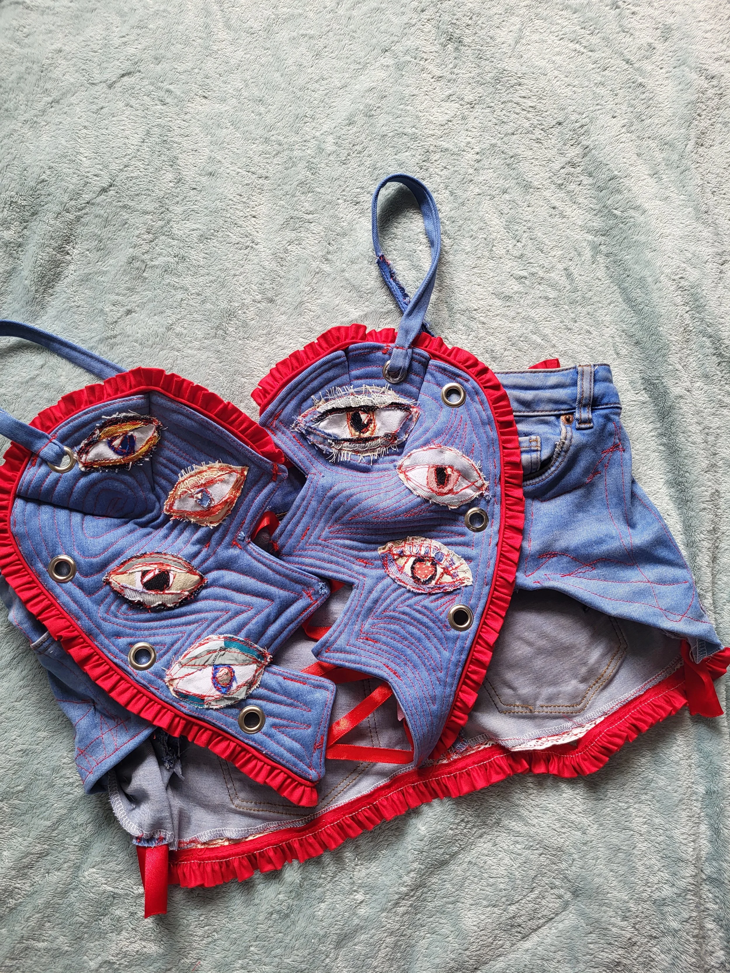 A denim vest and shorts decorated with embroidered eyes and red ruffled trim laid on a soft textured surface.