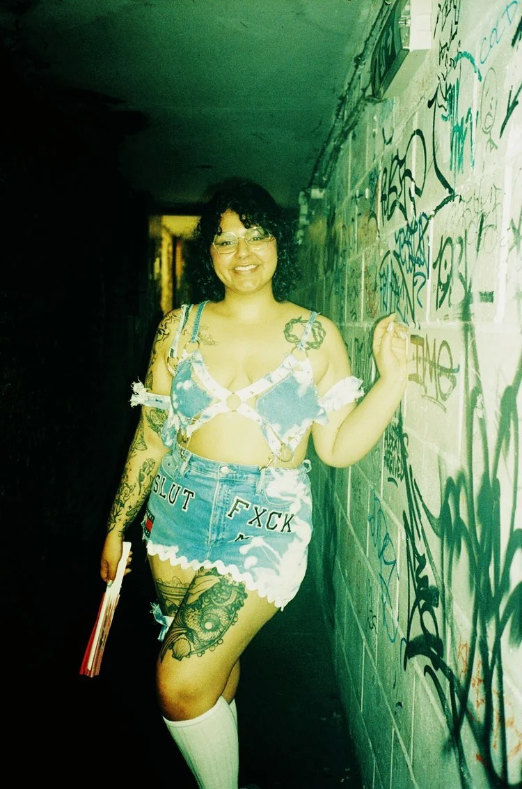A woman with glasses and tattoos standing against a graffiti-covered wall in a dimly lit alleyway, smiling at the camera.