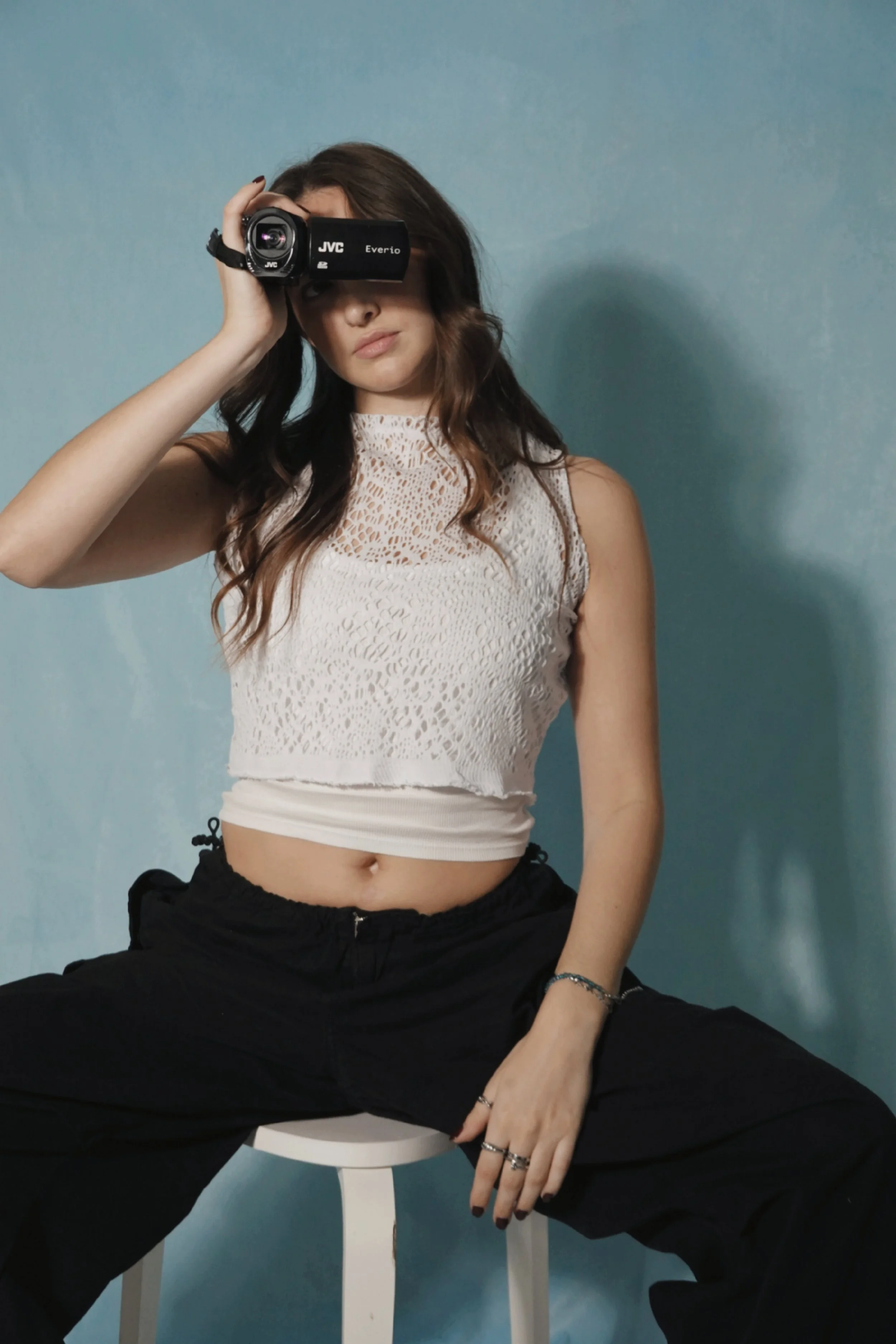 Young woman in white crochet crop top and black pants sits on a stool, holding a video camera to her eye, against a blue background.