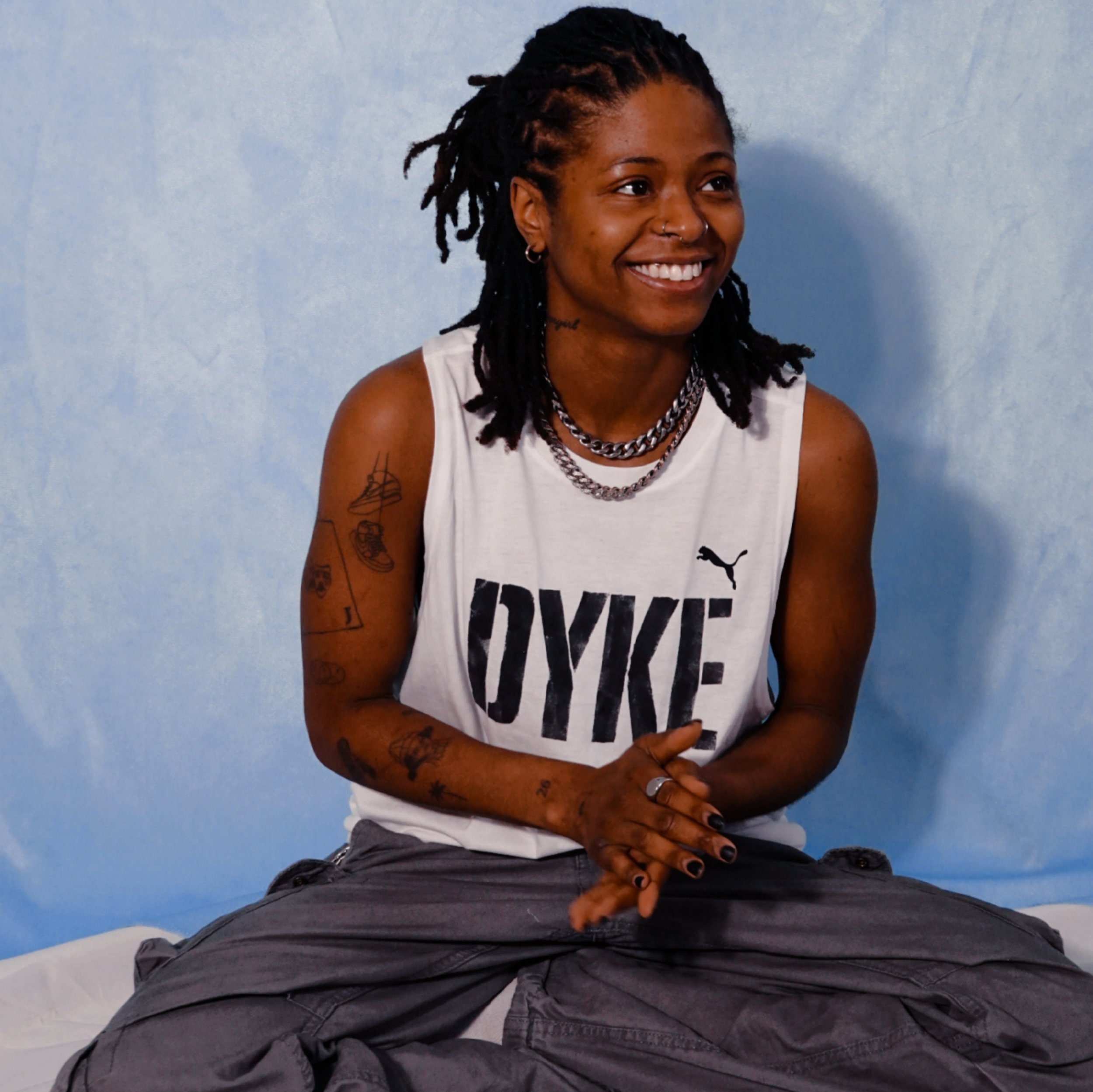 Smiling woman with locs, tattoos on her arms, wearing a white tank top with 'DYKE' and a Puma logo, sitting against a blue background.